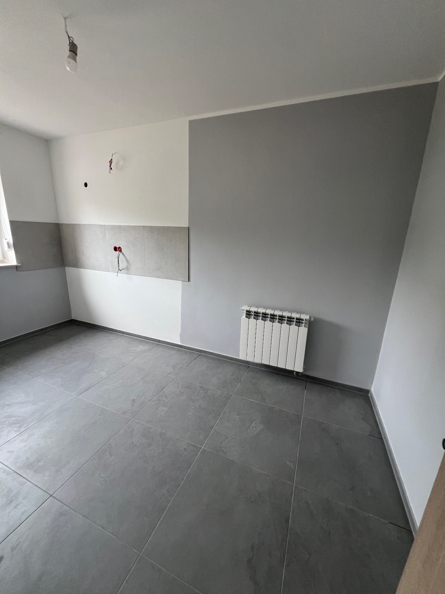 Wnętrze pokoju po częściowym remoncie: szare płytki na podłodze, fragment ściany pomalowany na szaro, grzejnik, widoczne przewody elektryczne.