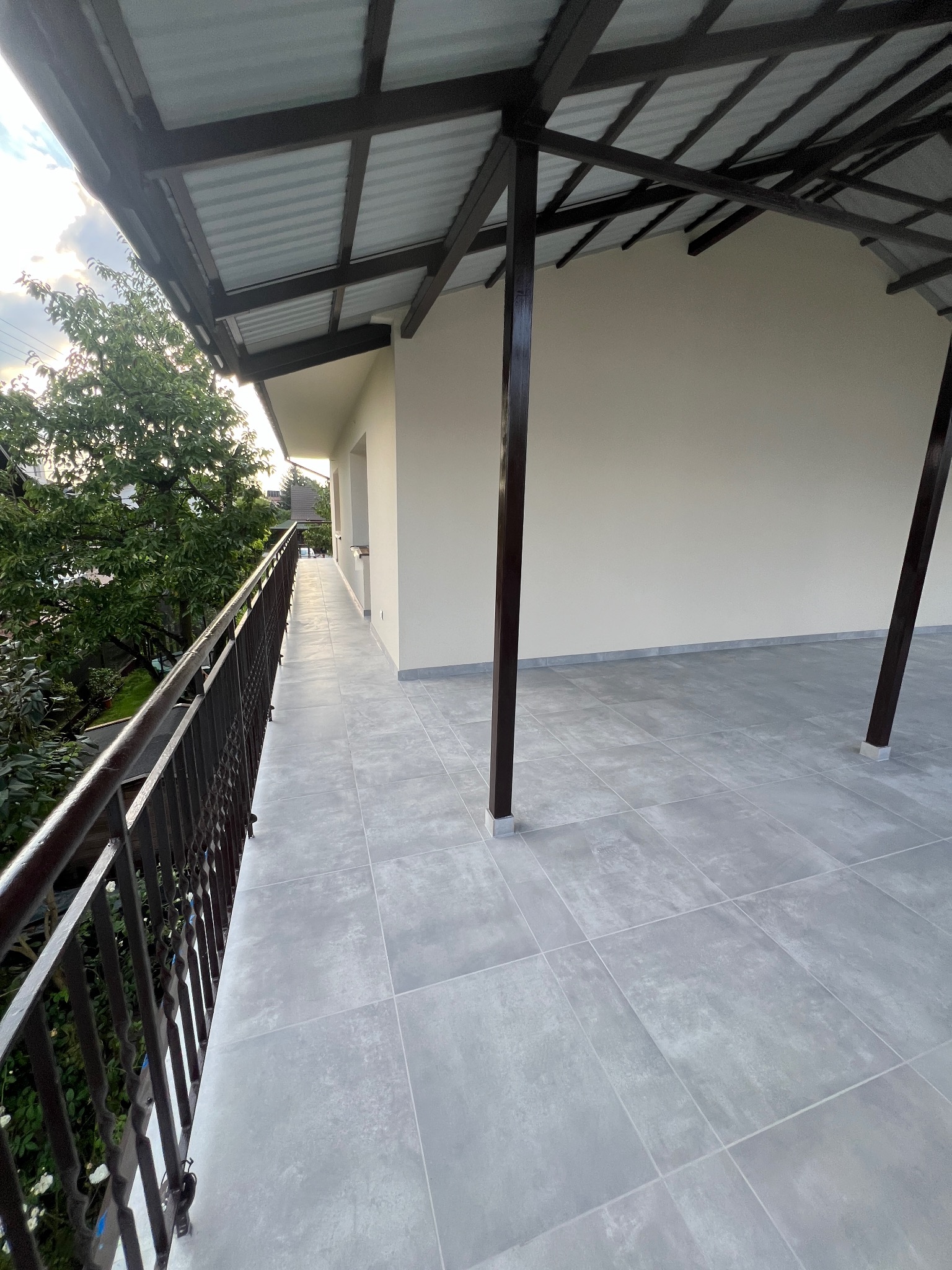 Długi, wykafelkowany balkon z metalową balustradą i zadaszeniem, świeżo po remoncie, widok na zieleń.