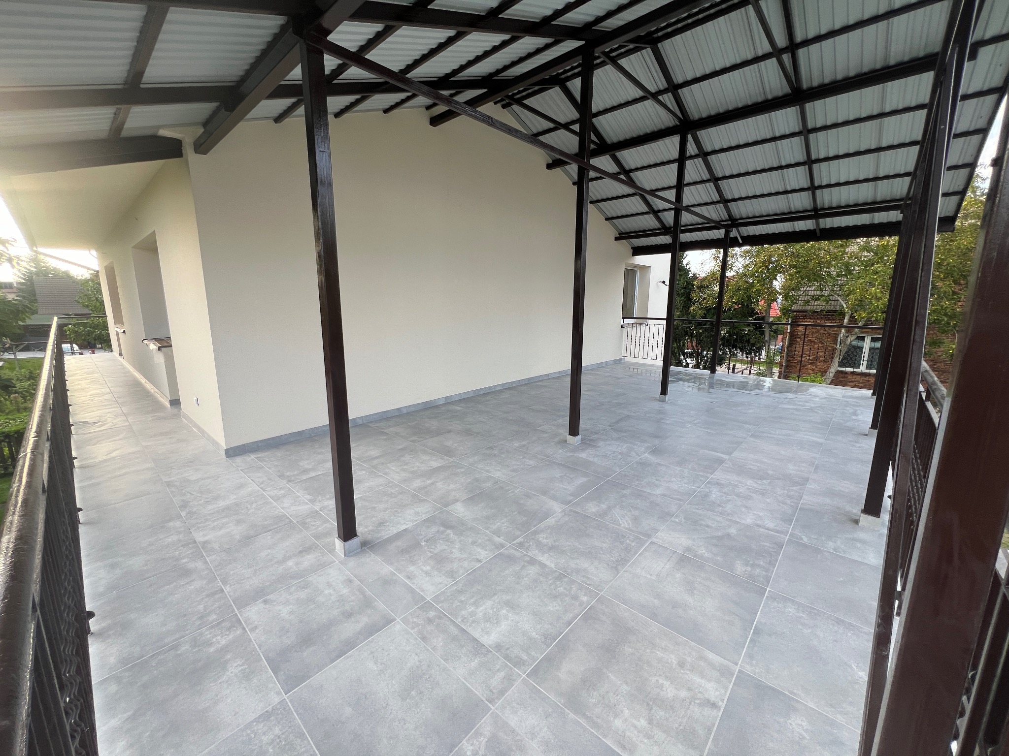 Zadaszony taras z szarymi płytkami podłogowymi ułożonymi w kwadraty, widok z balustrady, z widocznymi elementami konstrukcji dachu i fragmentami elewacji budynku w tle.