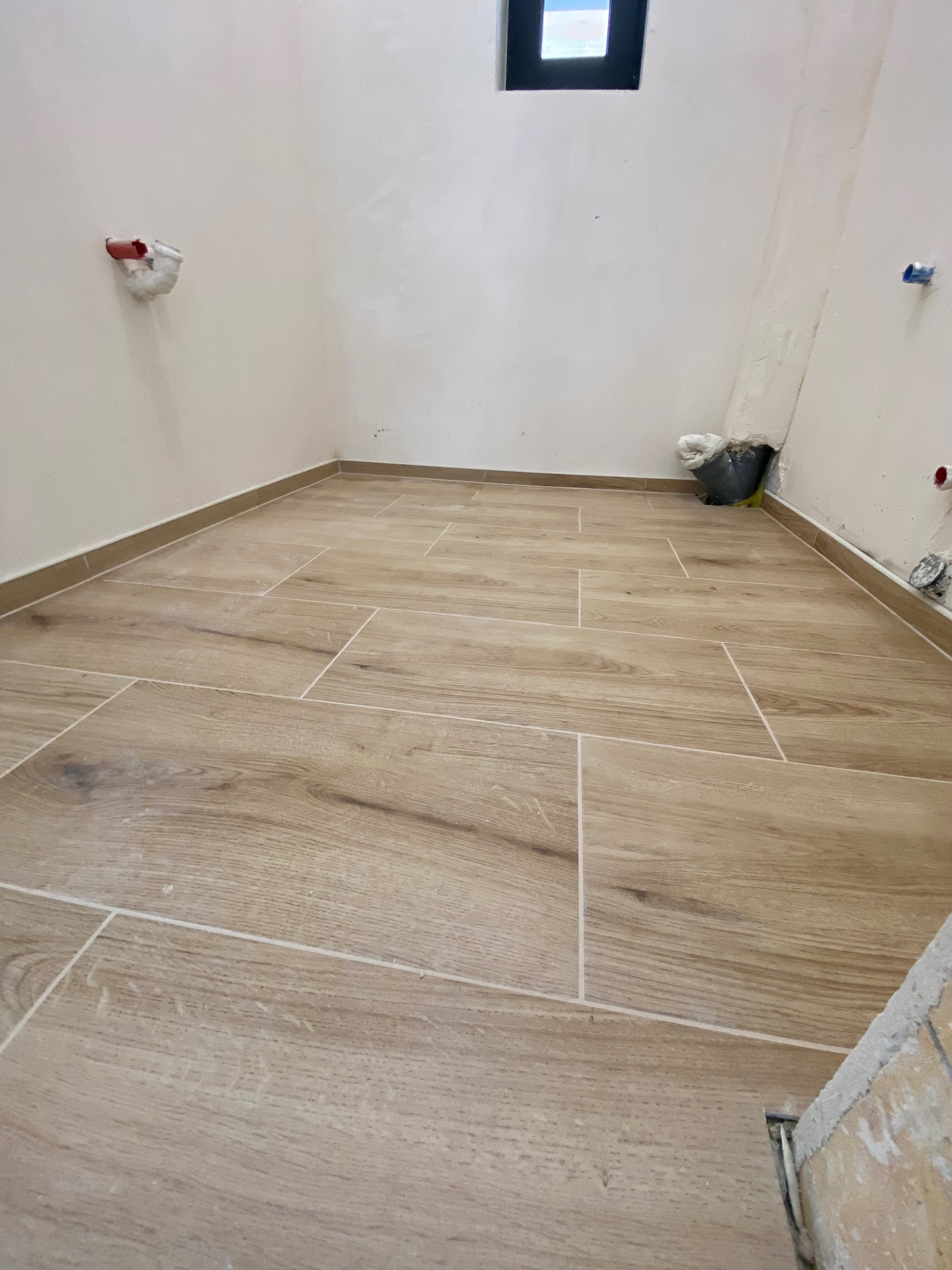 Ułożona podłoga z płytek imitujących drewno w jasnym odcieniu, widoczne fragmenty ścian z instalacjami, małe okno w górnej części ściany, wiadro z narzędziami w rogu pomieszczenia.