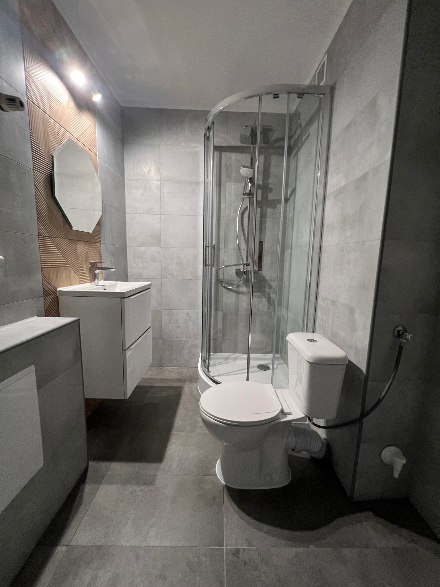 Nowoczesna łazienka z szarymi płytkami, kabiną prysznicową z zaokrąglonymi drzwiami, umywalką z szafką i dekoracyjną ścianą z drewnianymi elementami.