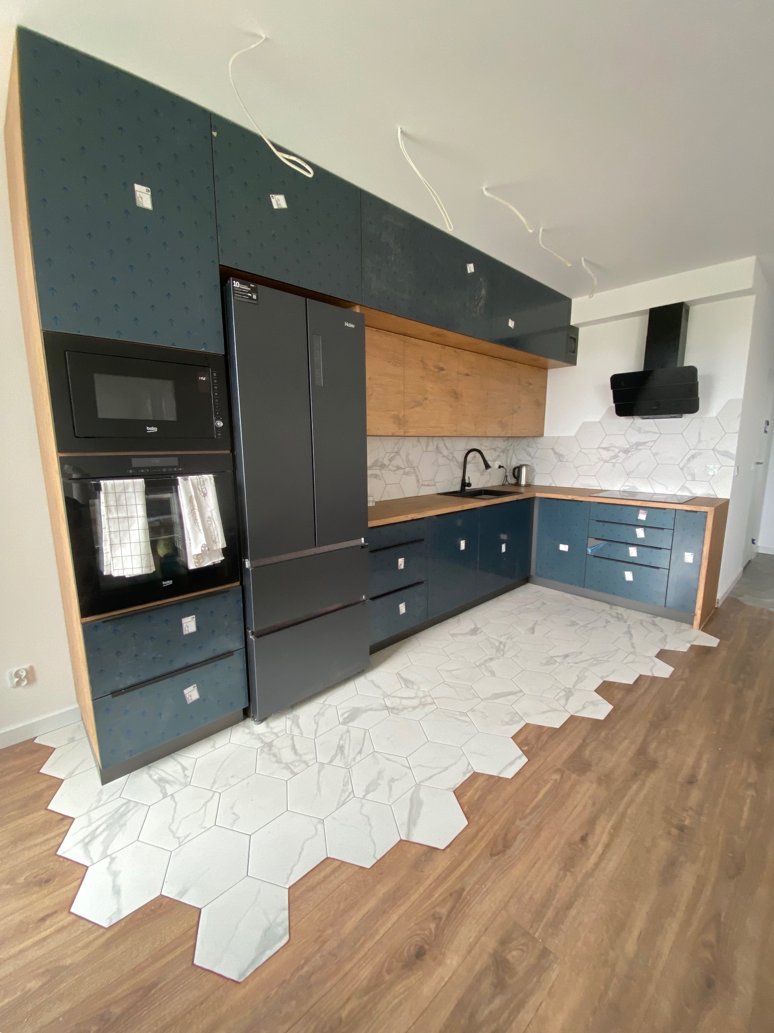 Niedokończona kuchnia z niebieskimi szafkami, drewnianymi blatami i heksagonalnymi płytkami podłogowymi przechodzącymi w panele, widoczne wiszące kable elektryczne.