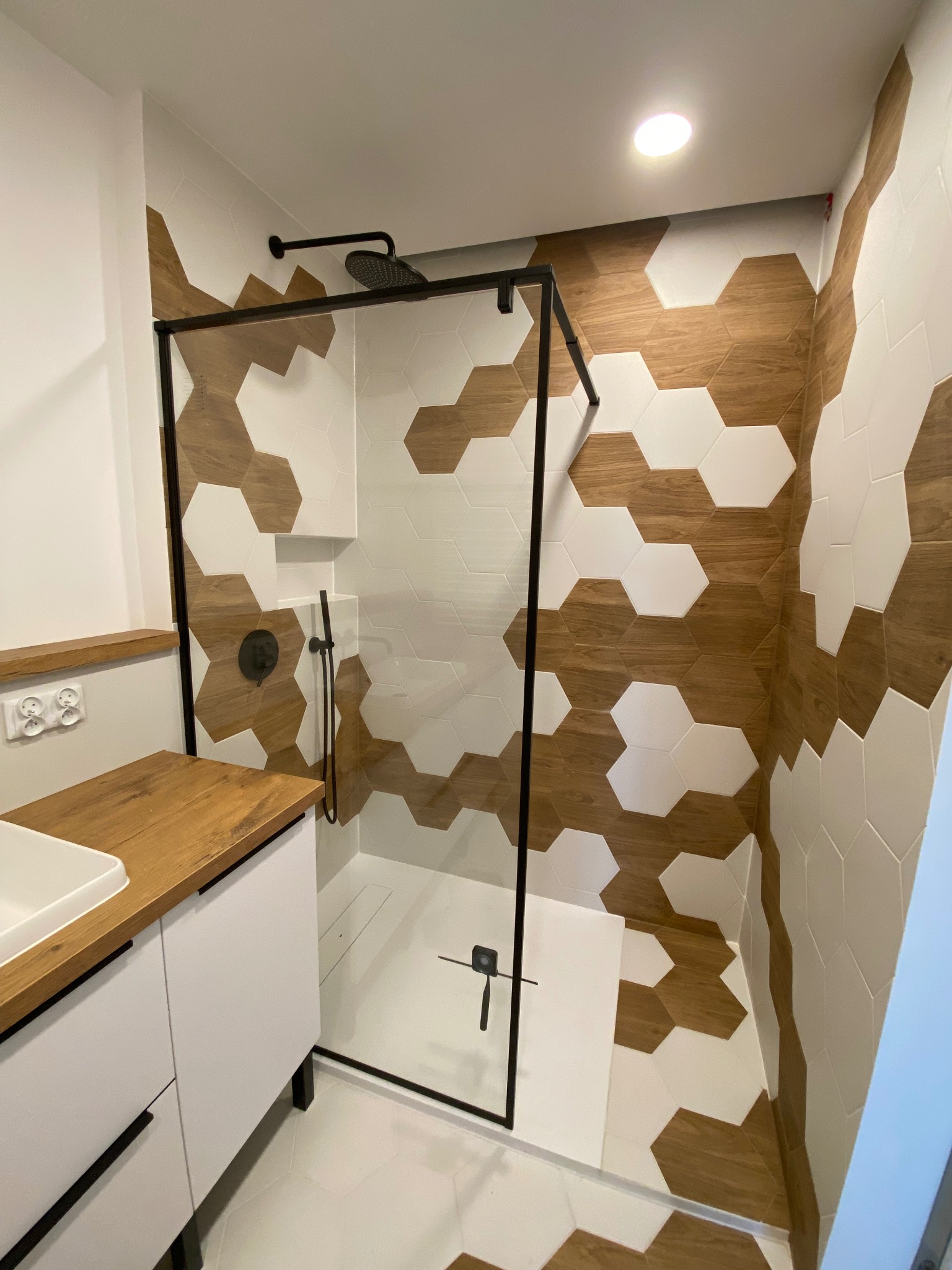 Nowoczesna łazienka z kabiną prysznicową typu walk-in, heksagonalne płytki w kolorze drewna i bieli, czarna armatura oraz biała szafka z drewnianym blatem.