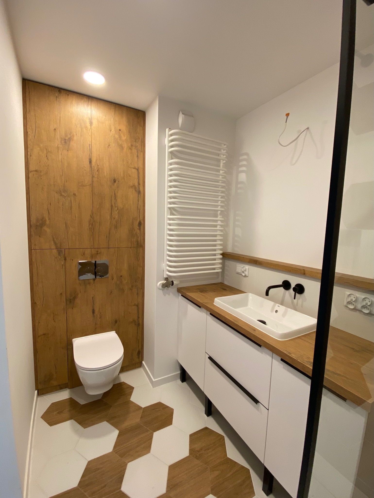 Nowoczesna łazienka z drewnianymi akcentami: zabudowa toalety z płyty imitującej drewno, heksagonalne płytki podłogowe w odcieniach bieli i drewna, biała szafka pod umywalkę z drewnianym blatem...
