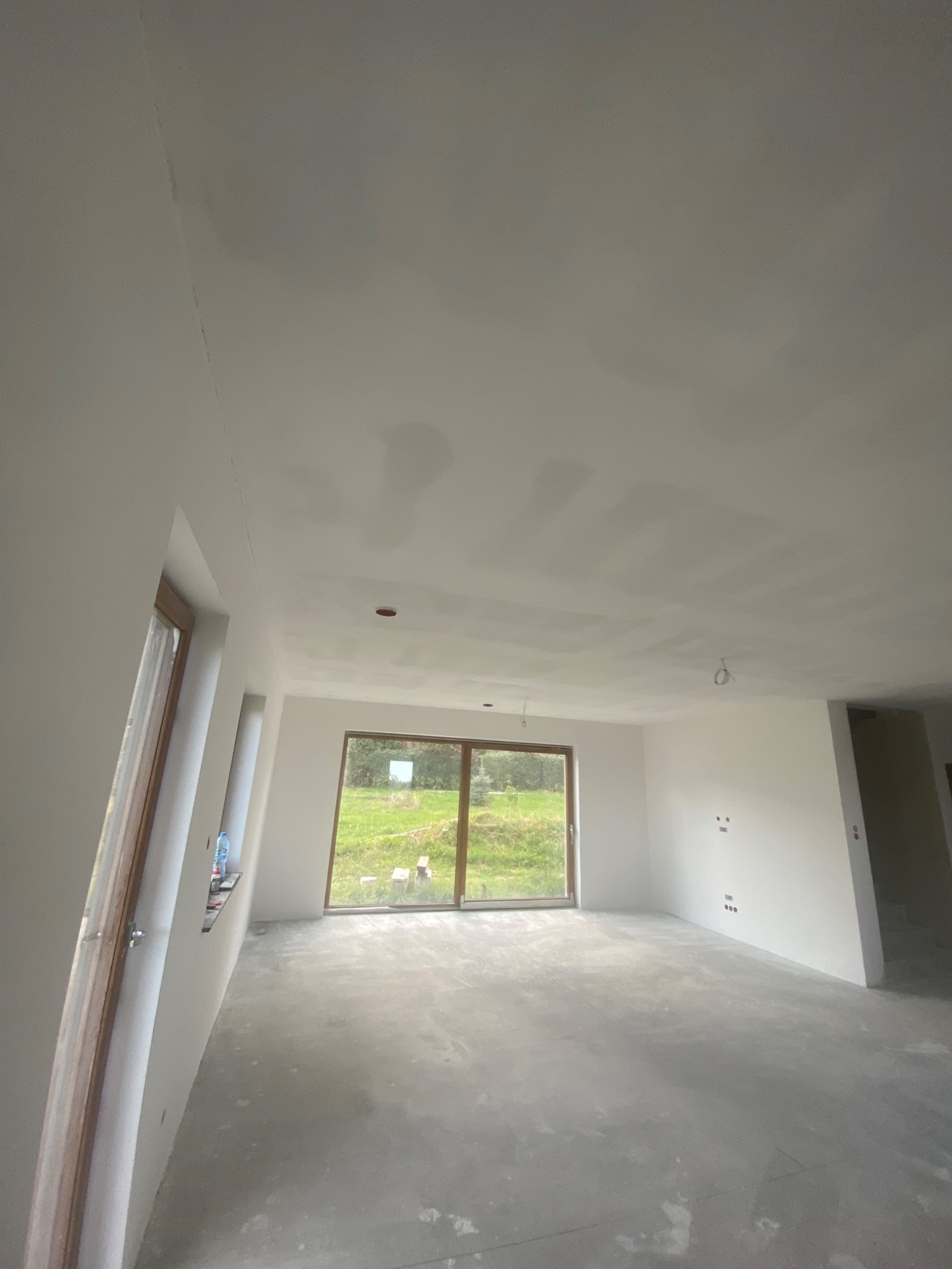 Wnętrze w trakcie prac wykończeniowych: sufit z widocznymi łączeniami płyt kartonowo-gipsowych przed szpachlowaniem, ściany przygotowane pod malowanie, surowa podłoga i duże okno z widokiem na zieleń.