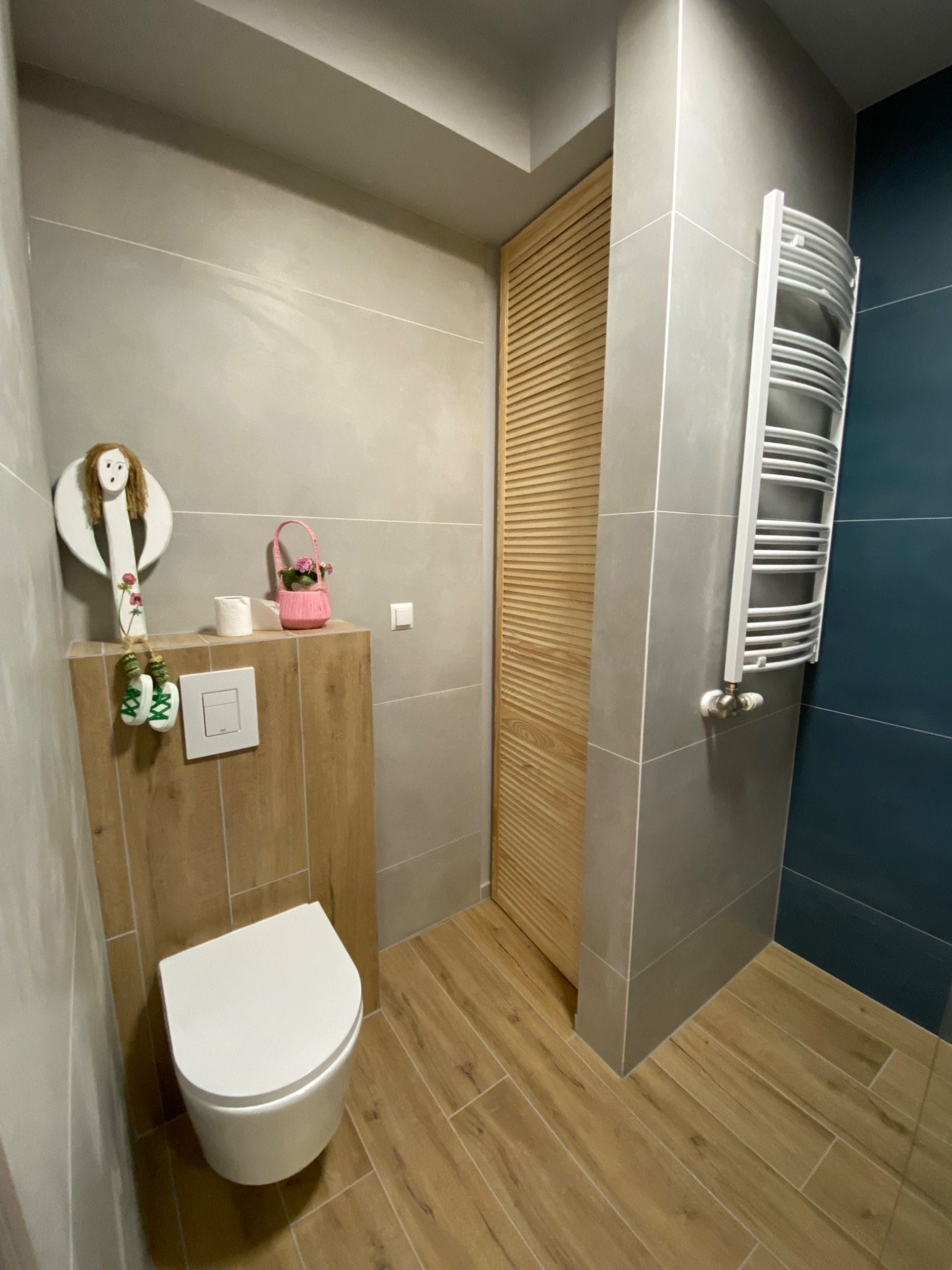 Nowoczesna łazienka z wiszącą toaletą, grzejnikiem drabinkowym i drewnianą podłogą imitującą panele, dekorowana ceramiczną figurką i różowym koszyczkiem z kwiatami.
