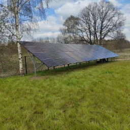 Instalacja fotowoltaiczna naziemna z panelami skierowanymi w stronę słońca, widoczna na tle trawnika, drzew i błękitnego nieba z chmurami.
