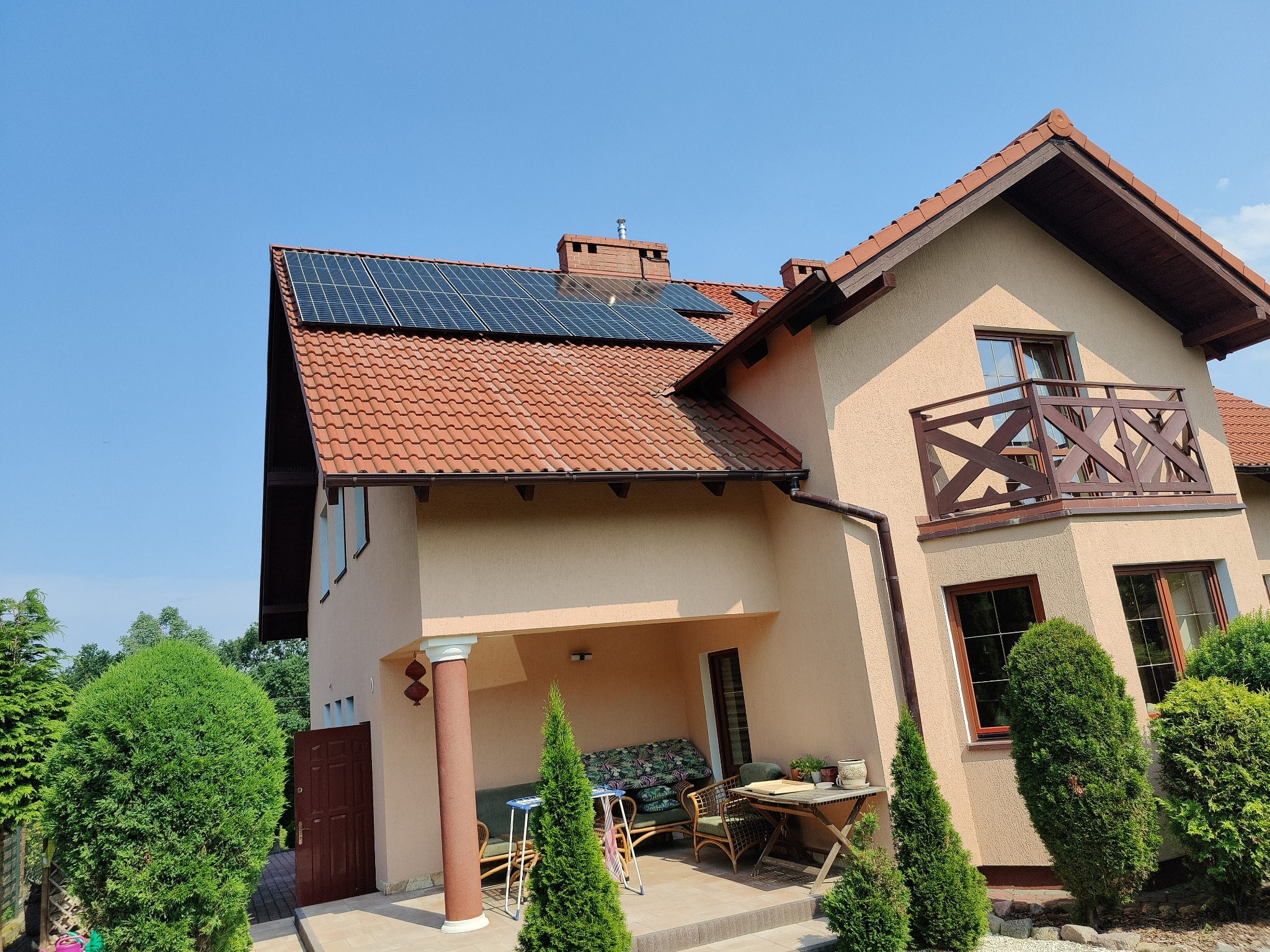 Widok z dołu na dom jednorodzinny z panelami słonecznymi na dachu pokrytym czerwoną dachówką, błękitne niebo w tle.