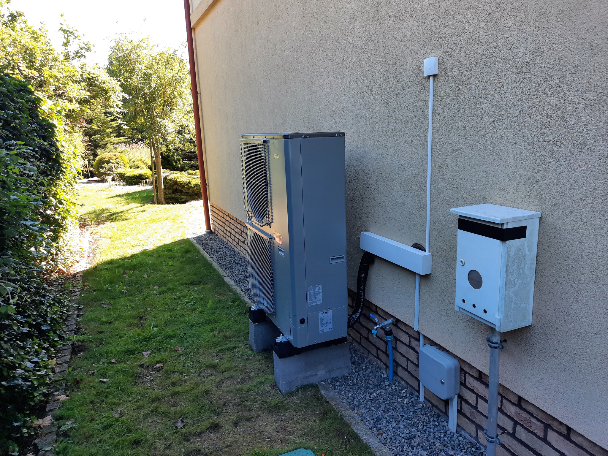 Zewnętrzna jednostka pompy ciepła NIBE ustawiona na betonowych blokach przy ścianie budynku z widoczną instalacją hydrauliczną i elektryczną.