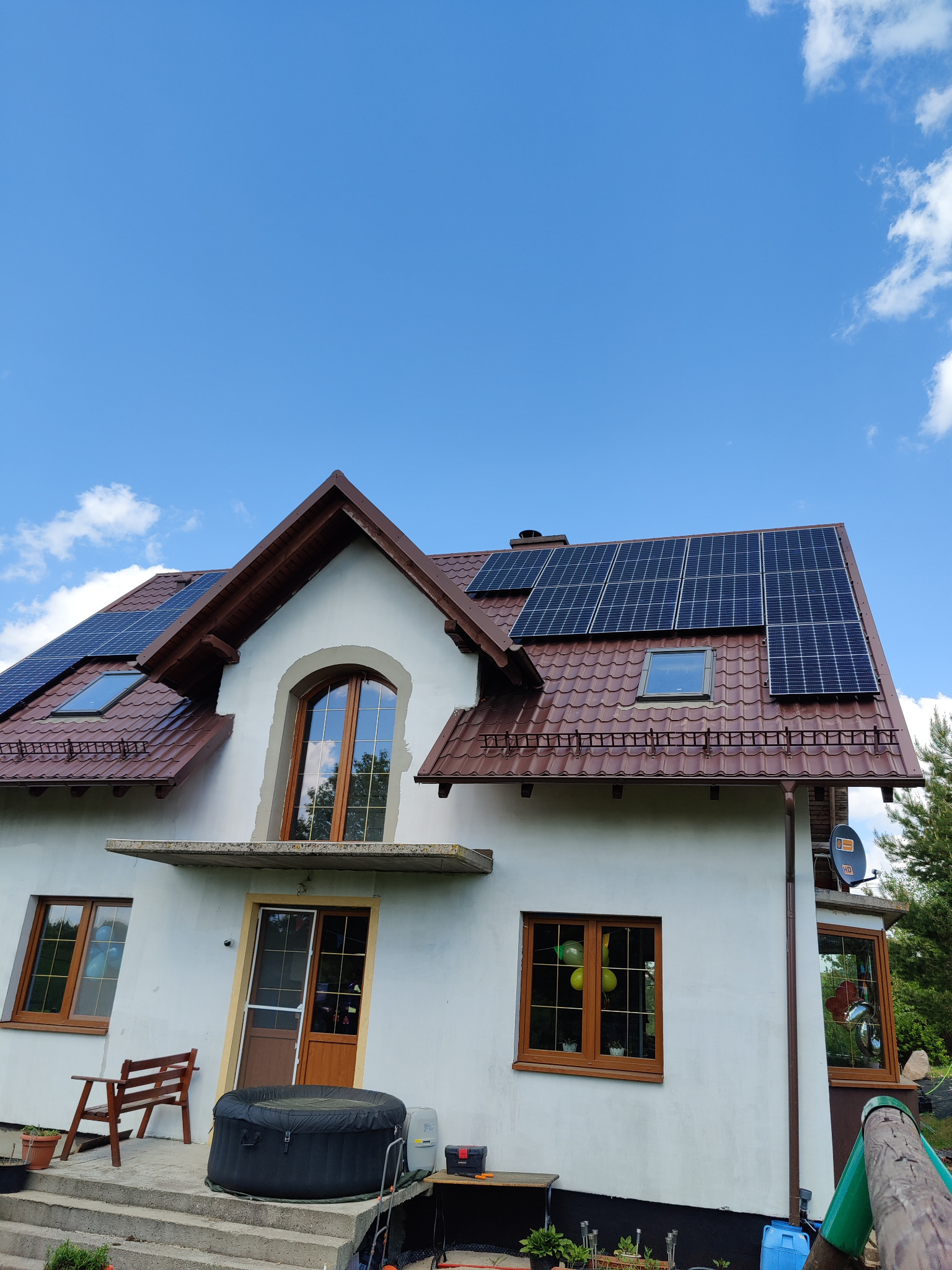 Dom jednorodzinny z brązowym dachem i panelami fotowoltaicznymi na połaci, widoczne okna dachowe, na tarasie jacuzzi i meble ogrodowe, błękitne niebo z chmurami.