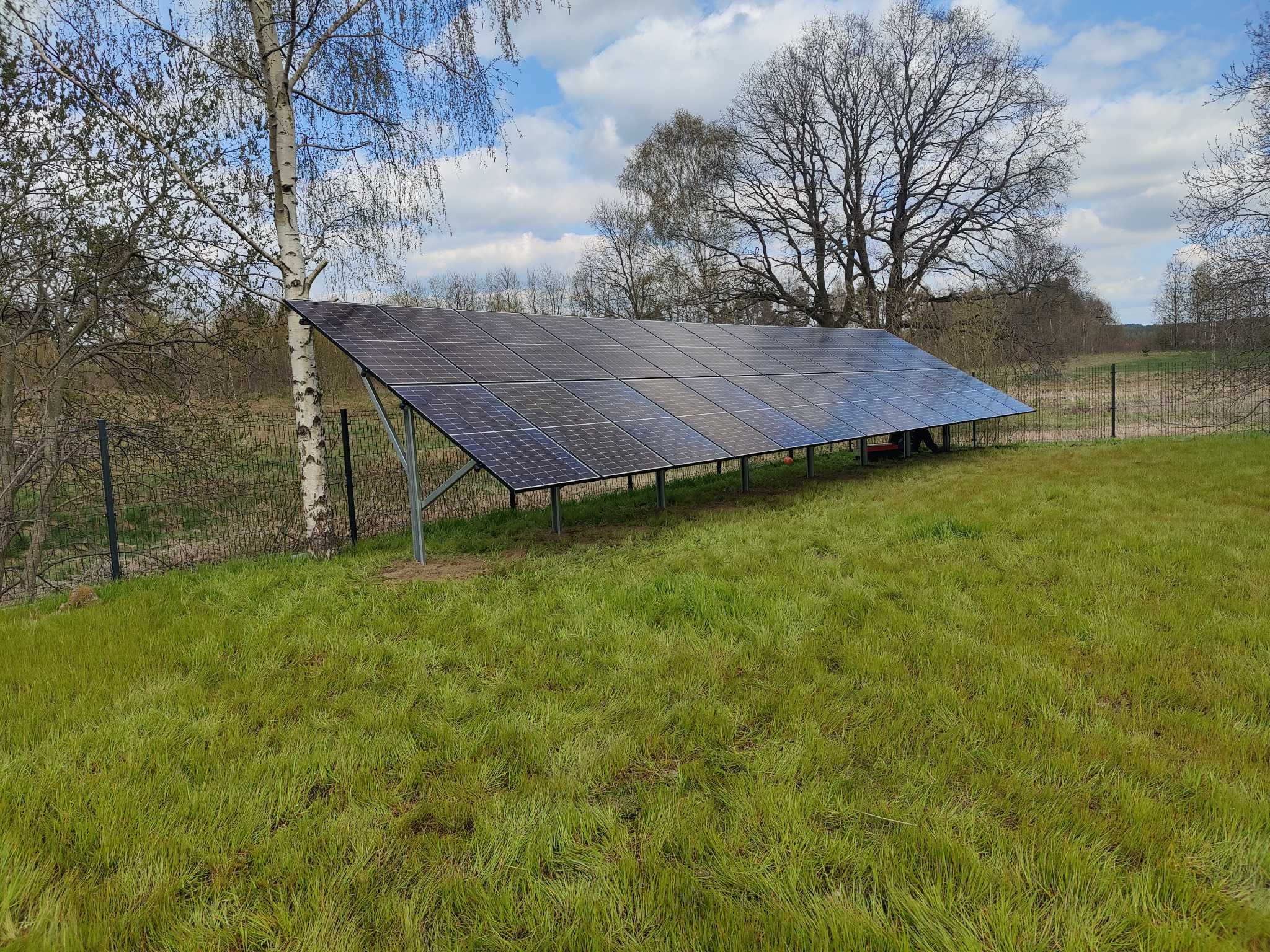 Instalacja fotowoltaiczna naziemna z panelami skierowanymi w stronę słońca, widoczna na tle trawnika, drzew i błękitnego nieba z chmurami.
