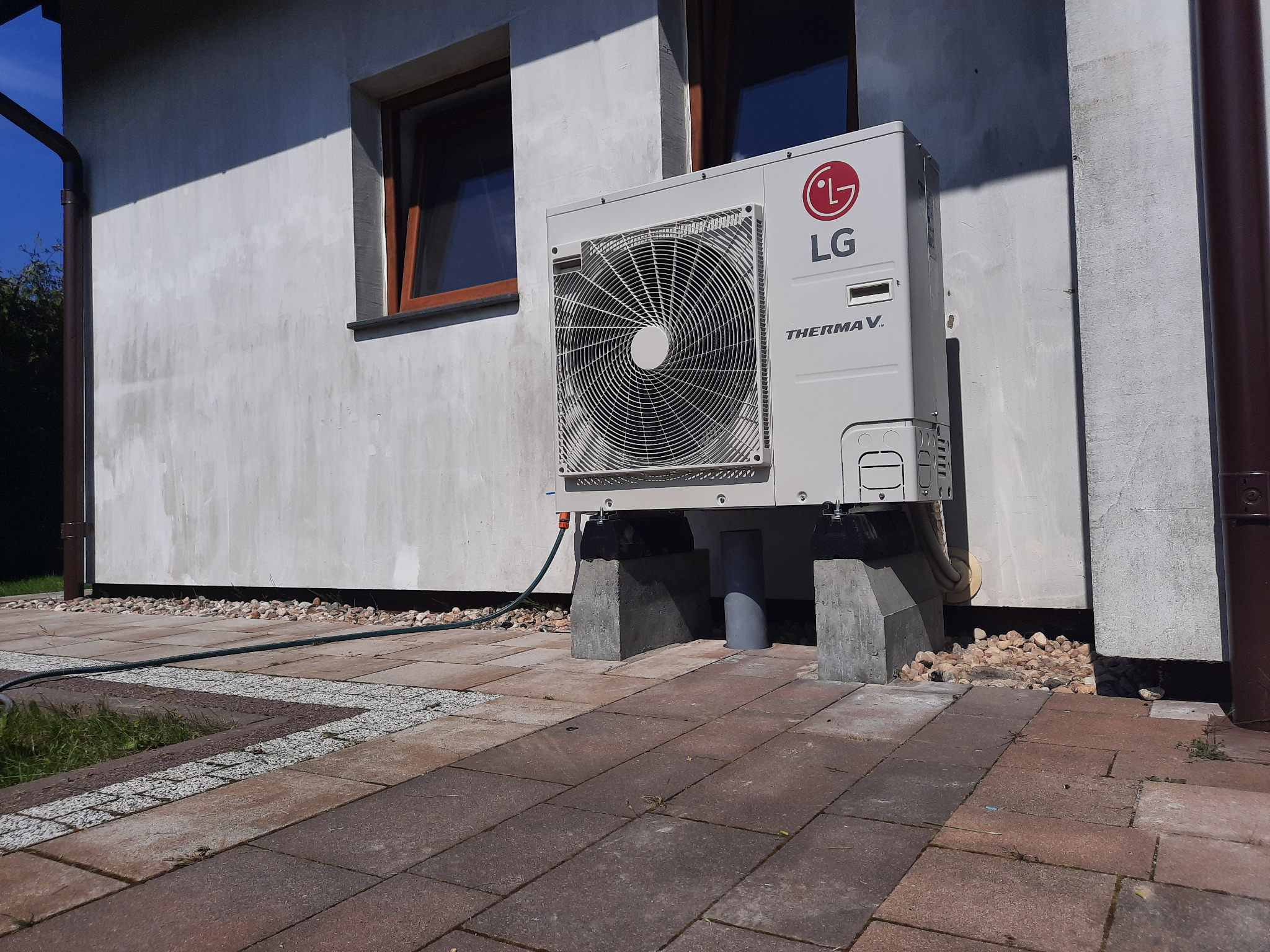 Biała pompa ciepła LG Therma V zamontowana na betonowych podstawach przy ścianie domu z widocznym oknem i rynną, otoczona brukowaną powierzchnią.