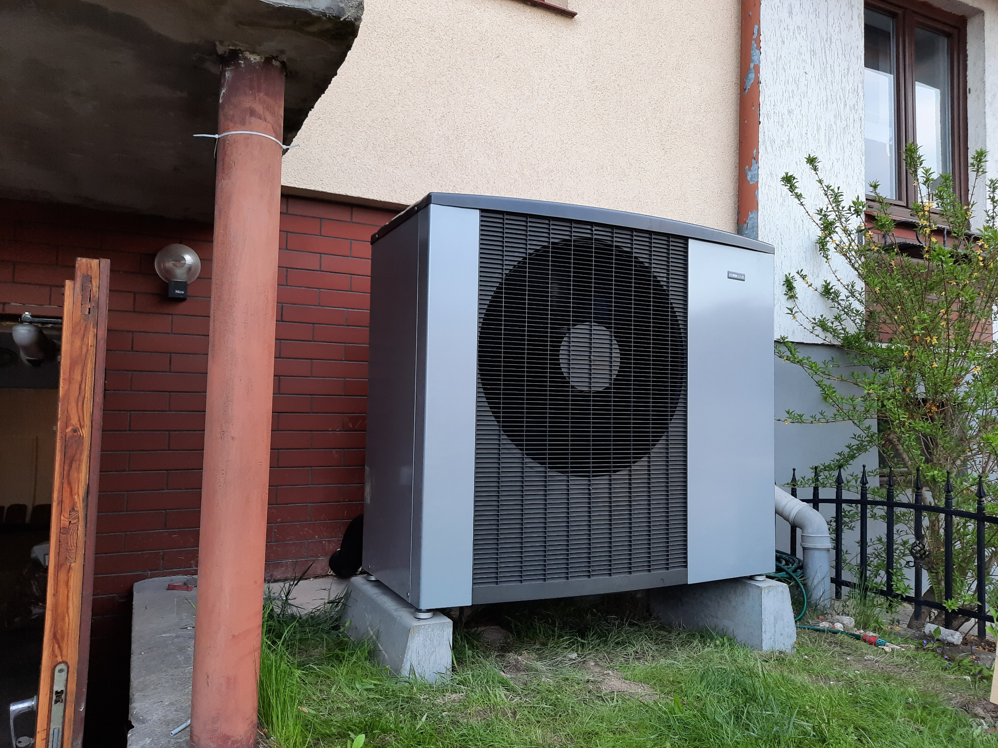 Szara pompa ciepła powietrze-woda na betonowych podstawach, umieszczona przy ścianie budynku z czerwoną cegłą i jasną elewacją, widoczny fragment czarnego ogrodzenia i roślinności.