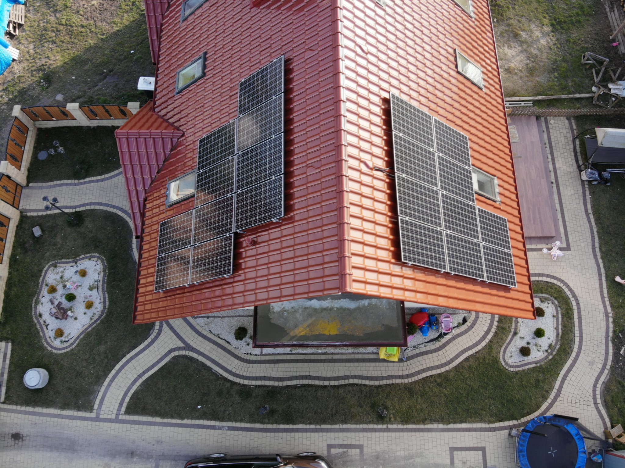 Widok z góry na dom z czerwoną dachówką i panelami fotowoltaicznymi, otoczony zadbanym ogrodem z kostki brukowej i trawnika, w tle widoczny samochód.