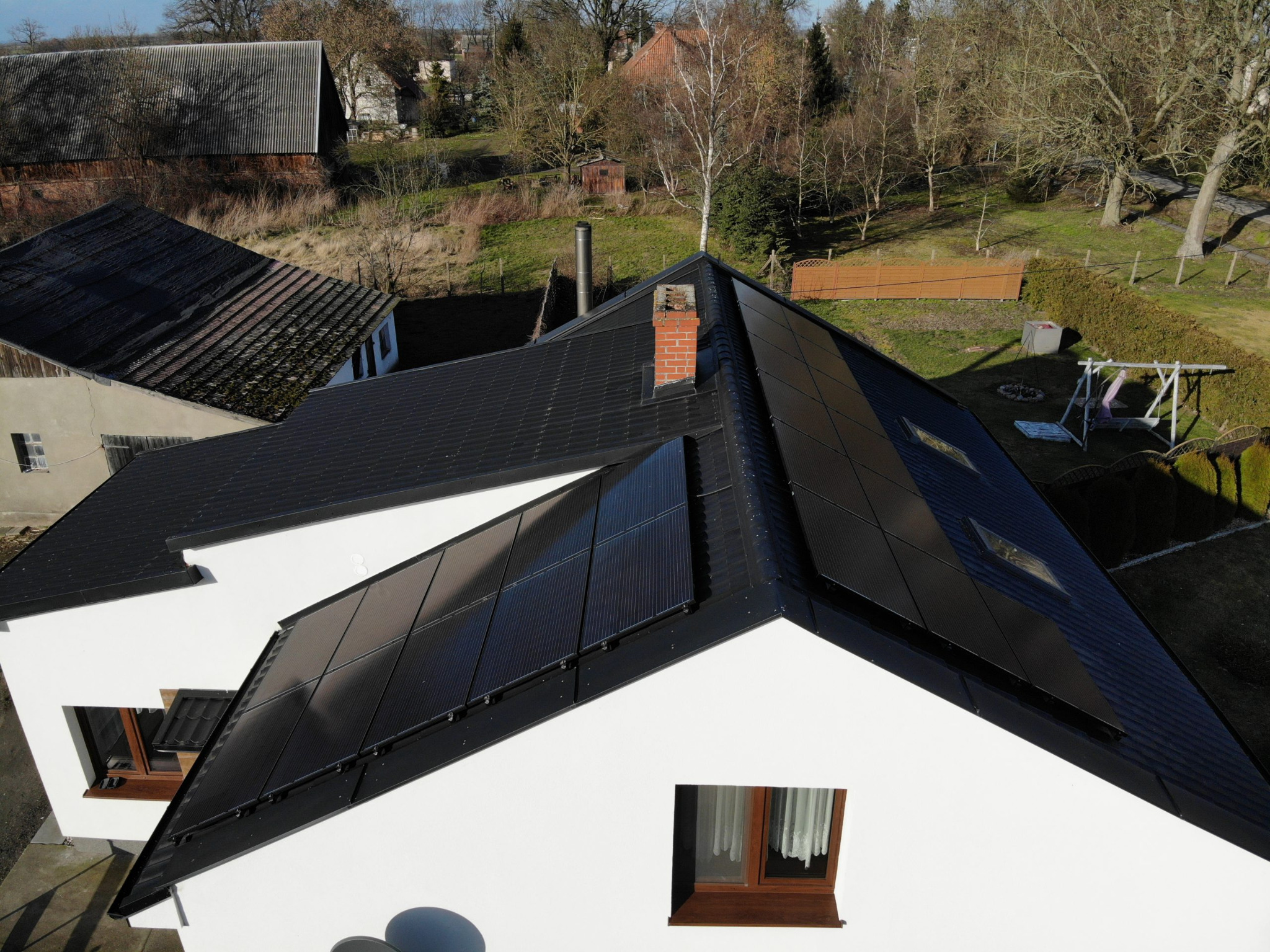 Widok z góry na czarne panele słoneczne zamontowane na dachu dwuspadowym białego domu jednorodzinnego z ceglanym kominem, w tle budynki gospodarcze i drzewa.