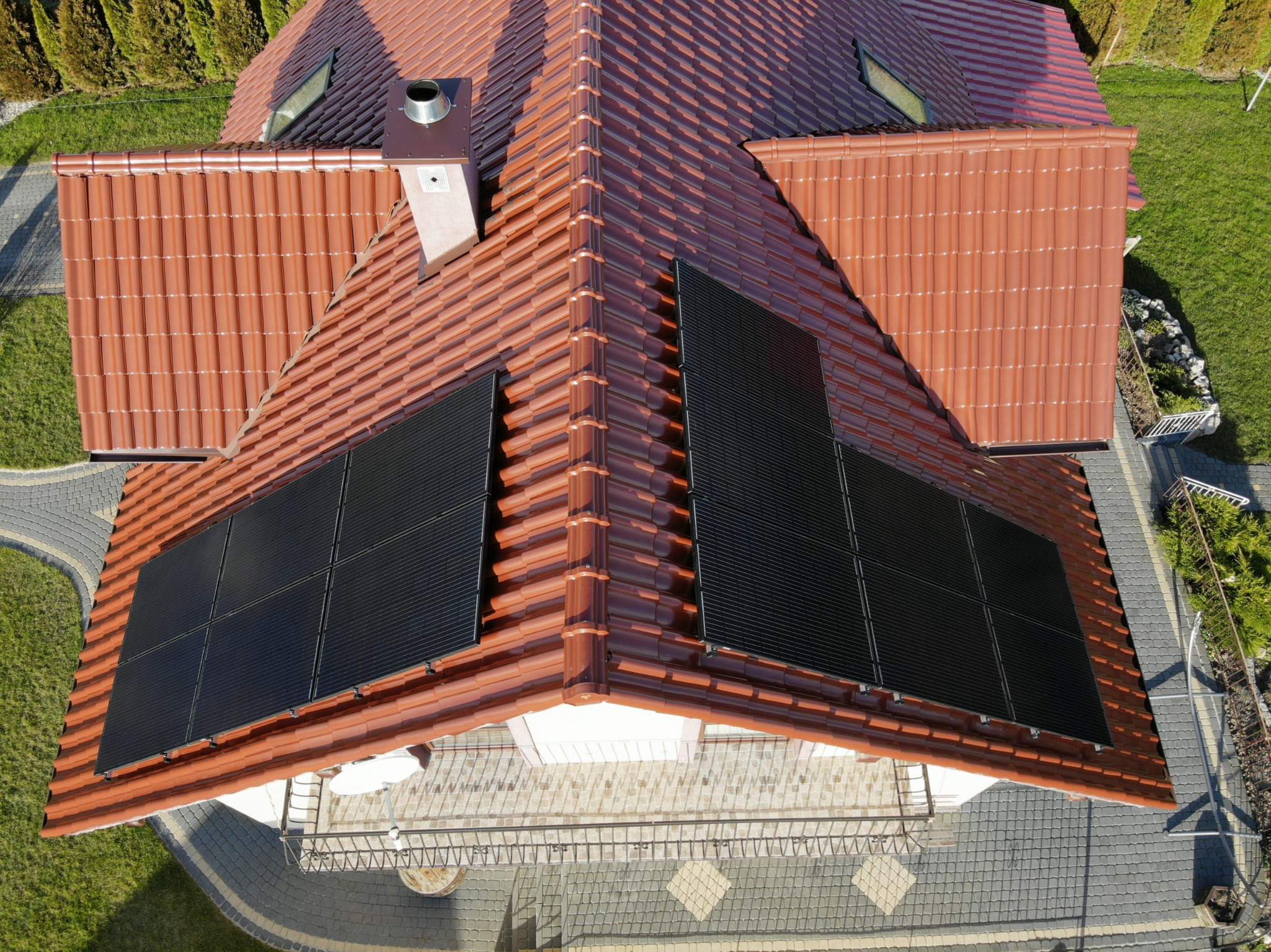 Widok z góry na dach domu z czerwoną dachówką i zamontowanymi czarnymi panelami słonecznymi, widoczny komin i otoczenie budynku z zielonym trawnikiem i kostką brukową.