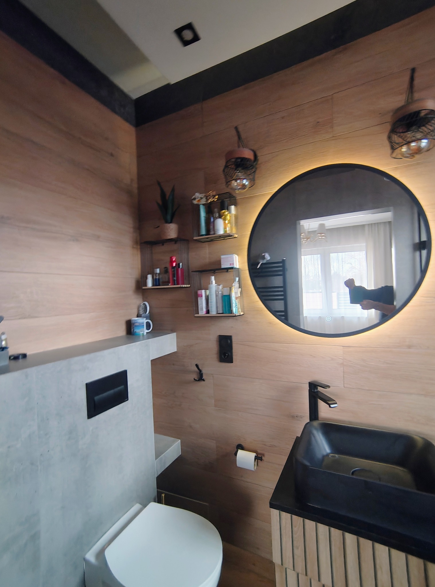 Nowoczesna łazienka z drewnopodobnymi płytkami, okrągłym lustrem z podświetleniem LED i czarną umywalką nablatową. Widoczne półki z kosmetykami i toaleta.