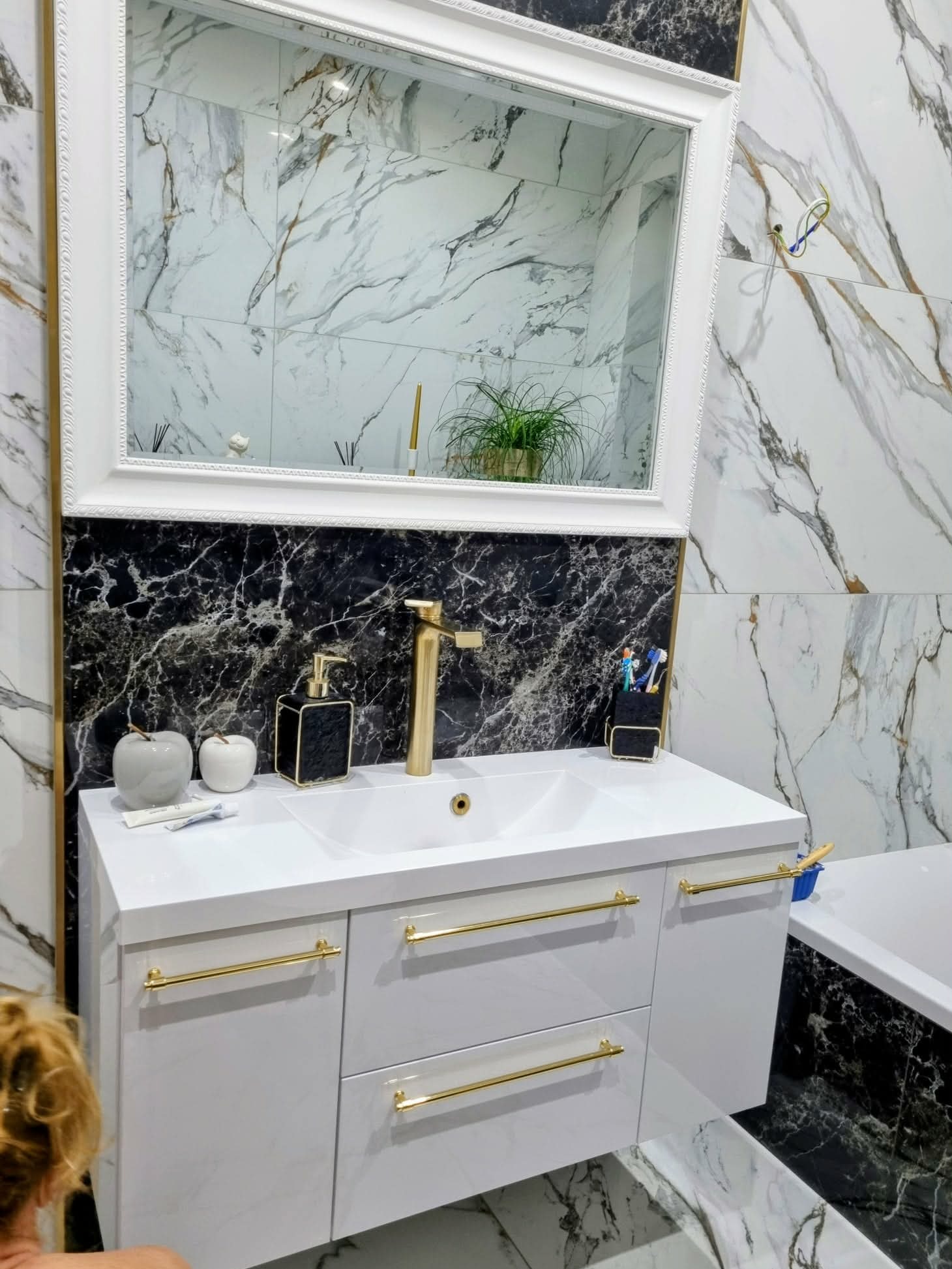 Elegancka łazienka z umywalką na szafce z białymi frontami i złotymi uchwytami. Marmurowe płytki na ścianach i podłodze. Nad umywalką lustro w białej ramie.