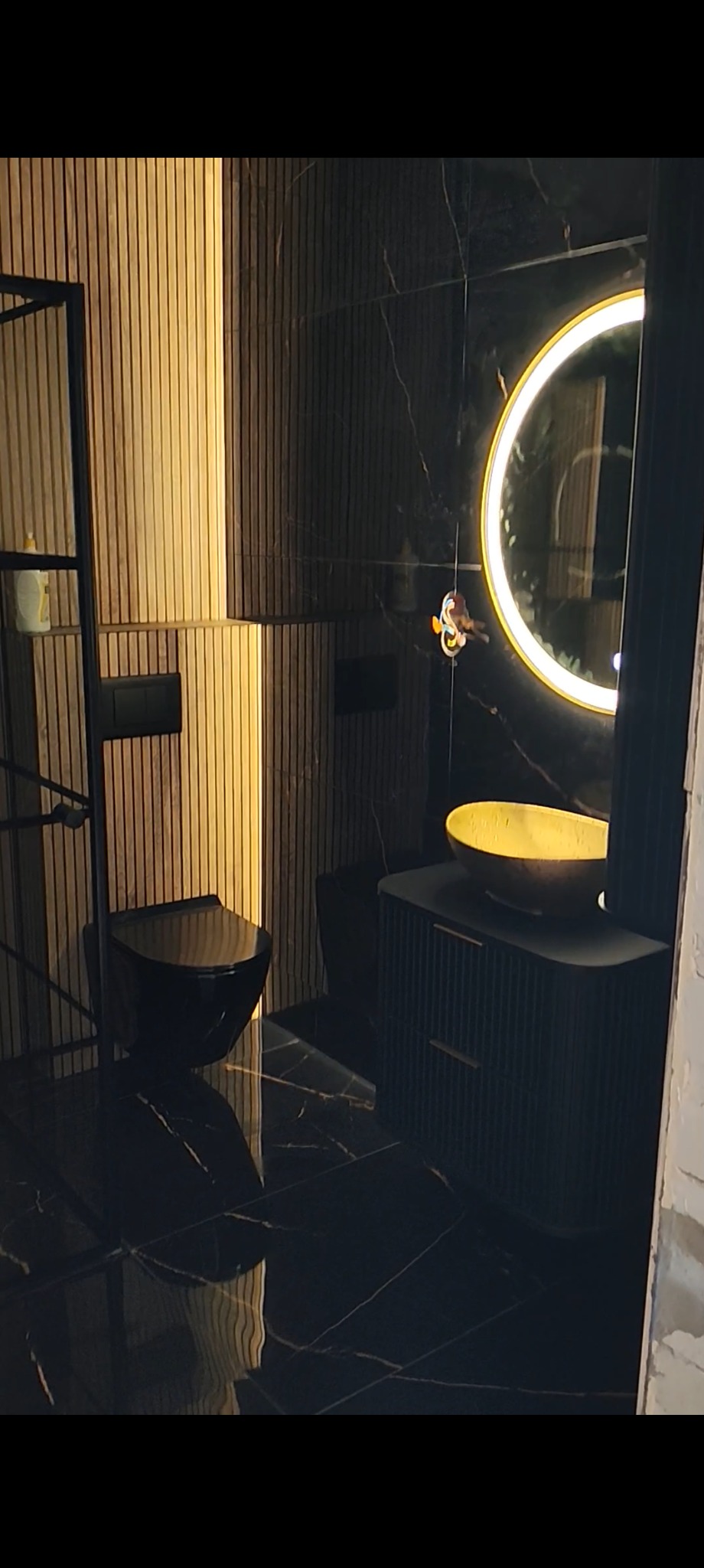 Nowoczesna łazienka z czarnymi płytkami imitującymi marmur, podświetlanym lustrem i elementami w kolorze złota. Uwagę zwraca wisząca toaleta i umywalka na szafce.