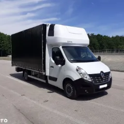 Biały Renault Master z czarną plandeką na parkingu, widok z przodu i z boku, błękitne niebo w tle.