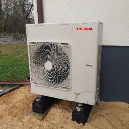 Zewnętrzna jednostka pompy ciepła Toshiba umieszczona na betonowych wspornikach w piasku, widoczny fragment elewacji budynku i rura spustowa.