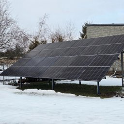 Doradca ds. OZE - fotowoltaika/pompy ciepła - Zimowy krajobraz z panelami fotowoltaicznymi na metalowej konstrukcji, z budynkiem z szarego pustaka w tle i resztkami śniegu na trawie.