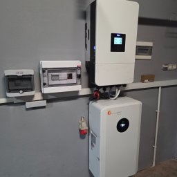 Doradca ds. OZE - fotowoltaika/pompy ciepła - Biały inwerter fotowoltaiczny Deye i magazyn energii Felicity Solar zamontowane na szarej ścianie wewnątrz budynku. Widoczne skrzynki elektryczne i gniazdo siłowe.