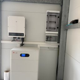 Doradca ds. OZE - fotowoltaika/pompy ciepła - Biały magazyn energii Felicity ESS z inwerterem i rozdzielnicą na ścianie z paneli. Widoczne kable i przewody. Instalacja w pomieszczeniu z metalową konstrukcją.