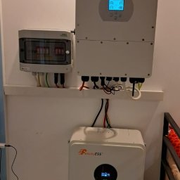 Doradca ds. OZE - fotowoltaika/pompy ciepła - Biały inwerter Deye i magazyn energii Felicity ESS zamontowane na ścianie, z widoczną skrzynką bezpiecznikową i okablowaniem. Instalacja wewnątrz budynku.