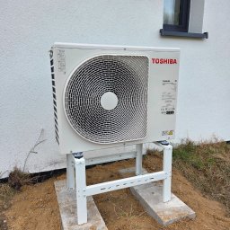 Doradca ds. OZE - fotowoltaika/pompy ciepła - Biała pompa ciepła Toshiba na metalowym stelażu, ustawiona na betonowych płytach przed jasnym budynkiem z małym oknem. Widoczne elementy instalacji i otoczenie.