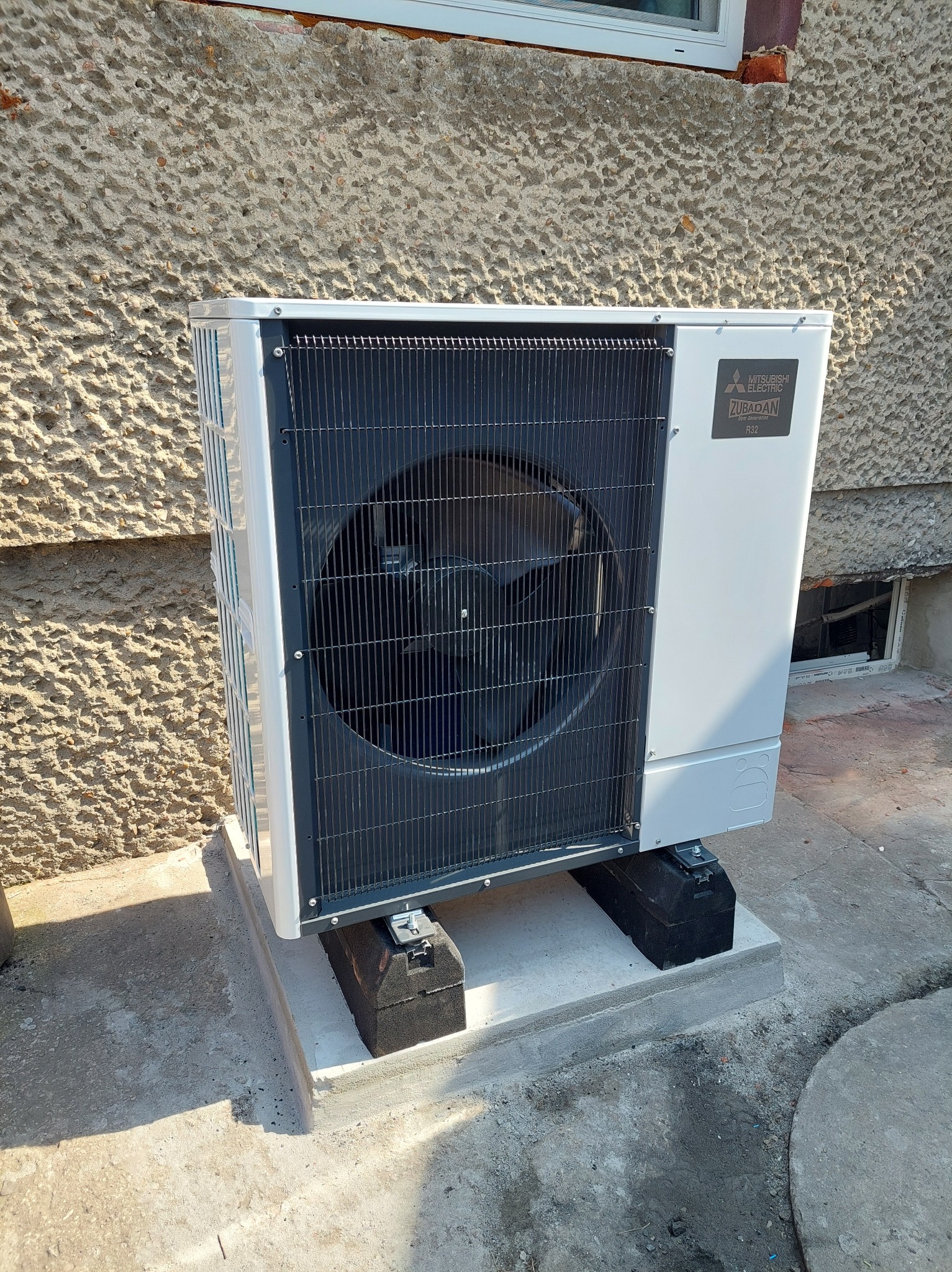 Biała jednostka zewnętrzna z czarną kratką wentylacyjną, marki Mitsubishi Electric Zubadan R32, zamontowana na betonowym fundamencie przed szorstką ścianą budynku.