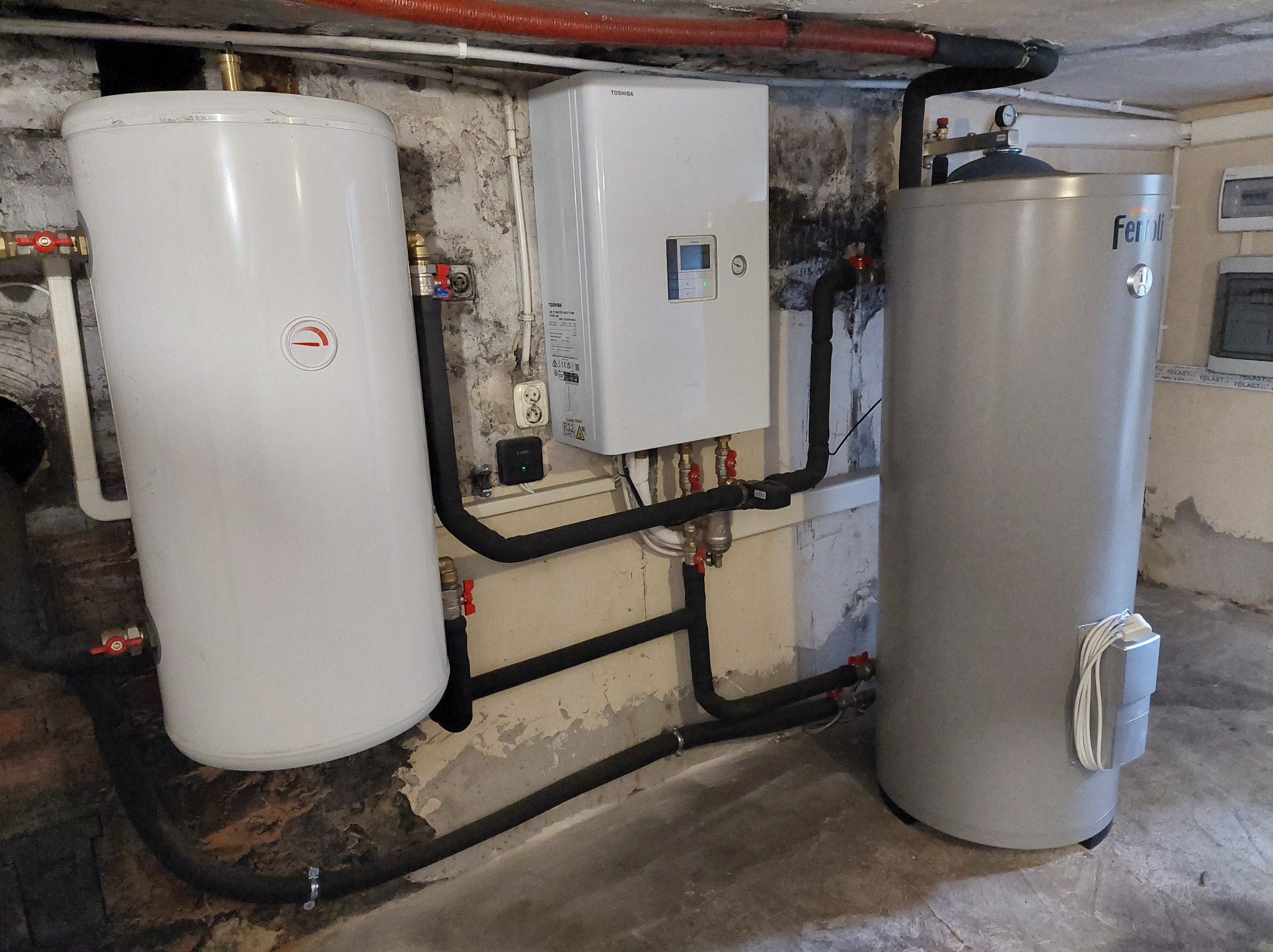 Instalacja w piwnicy z białym i szarym zbiornikiem oraz pompą ciepła Toshiba, widoczne rury i zawory.