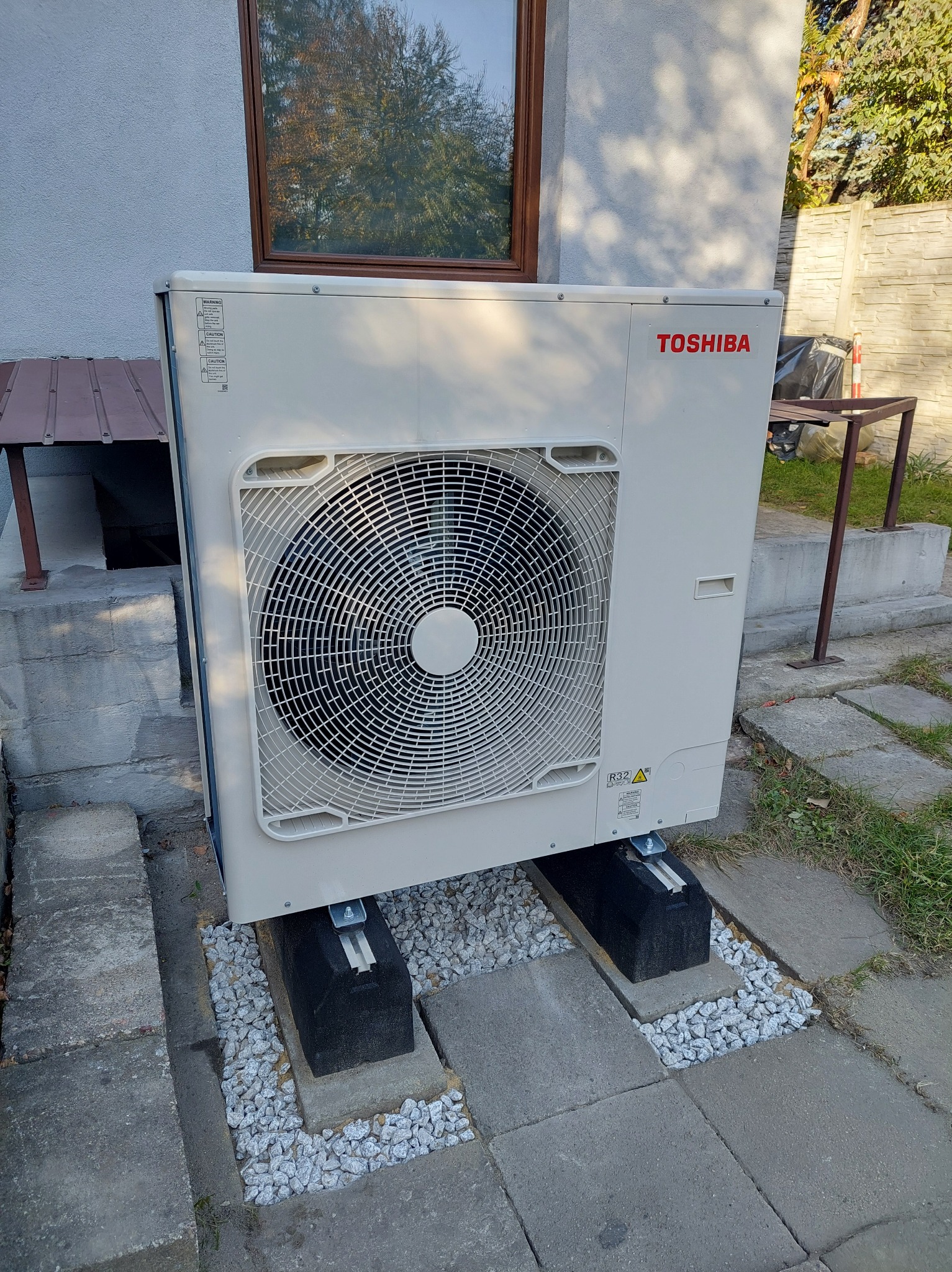 Zewnętrzna jednostka pompy ciepła Toshiba zamontowana na betonowych płytach z podsypką z białego żwiru, widoczna na tle elewacji domu i fragmentu ogrodu.
