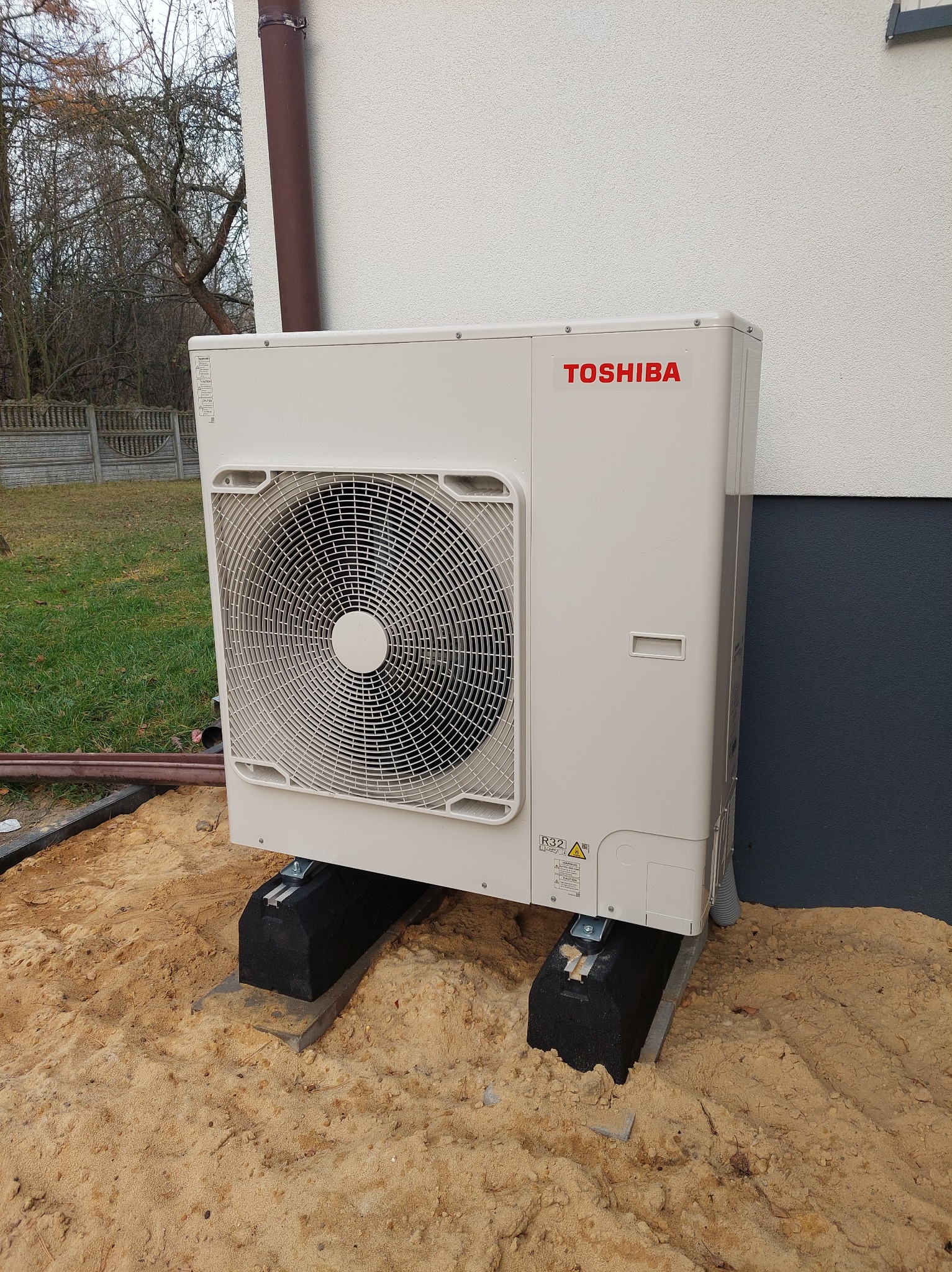 Zewnętrzna jednostka pompy ciepła Toshiba umieszczona na betonowych wspornikach w piasku, widoczny fragment elewacji budynku i rura spustowa.