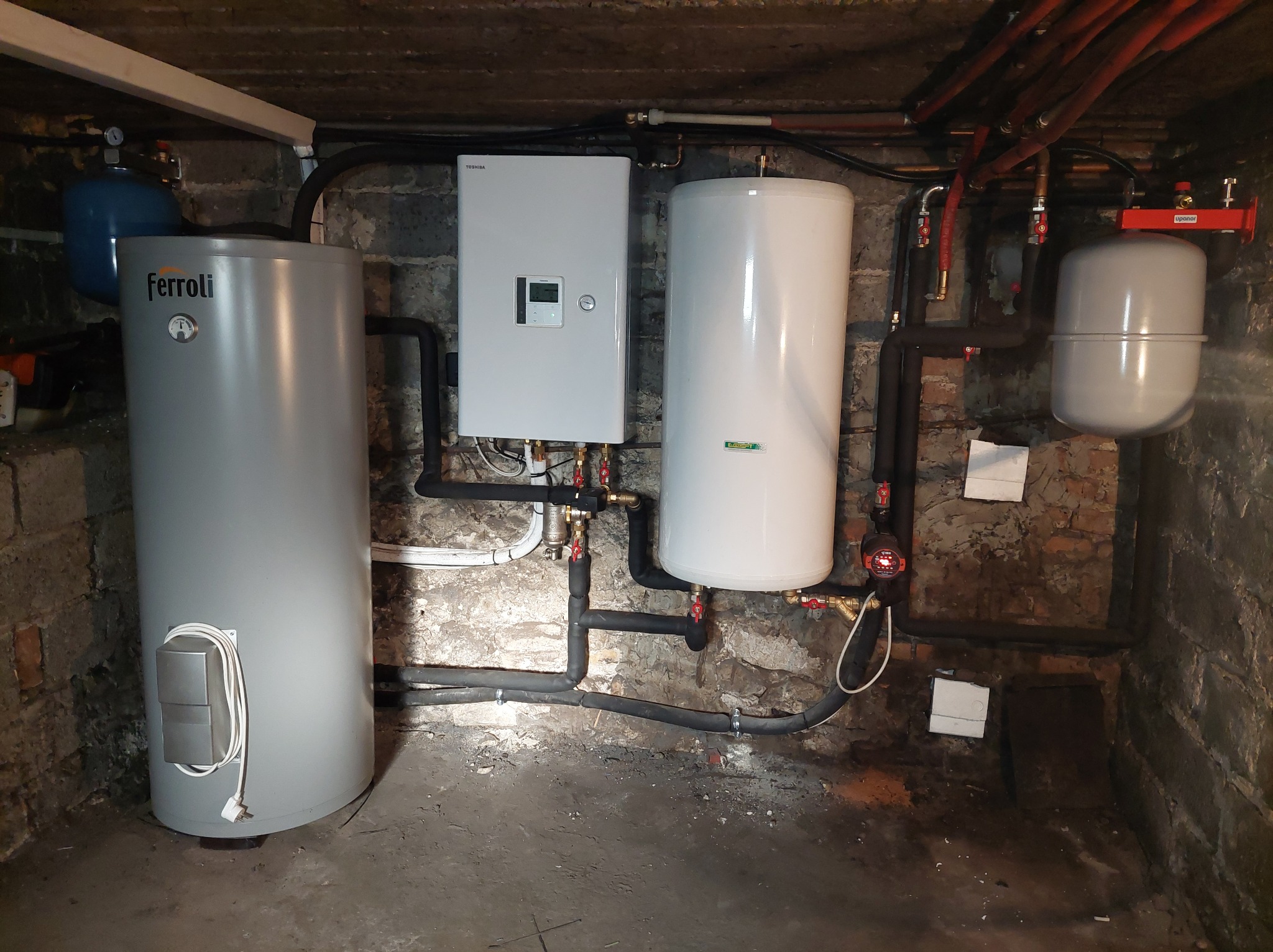 Instalacja grzewcza w piwnicy z kotłem Toshiba, zasobnikiem Ferroli i zbiornikami wyrównawczymi UpaNer, rury w izolacji, surowa ściana z cegły.