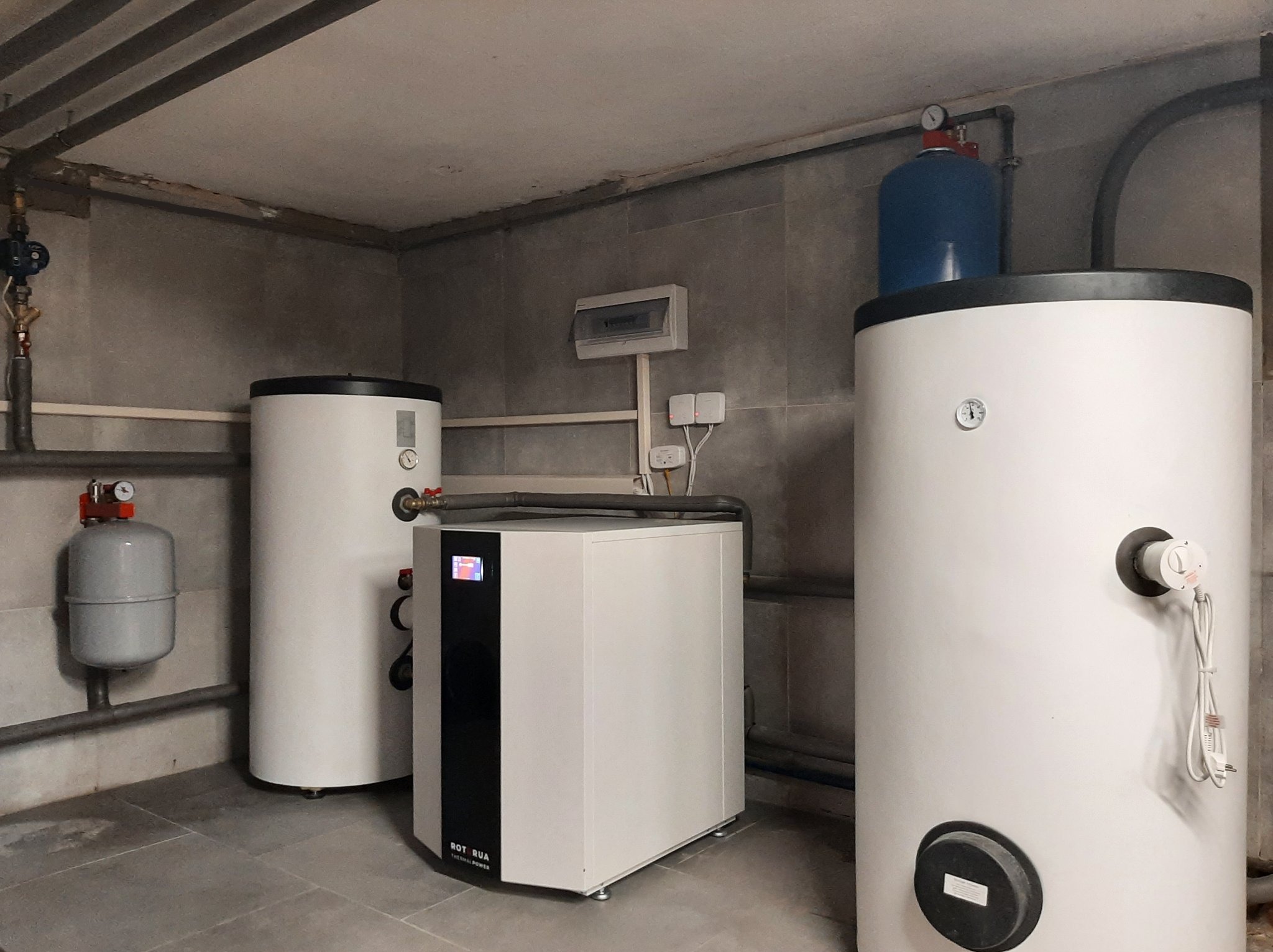 Instalacja w kotłowni: Dwa białe, cylindryczne zasobniki ciepłej wody oraz biała pompa ciepła typu monoblok z czarnym panelem przednim, na szarych płytkach podłogowych i tle szarych ścian.