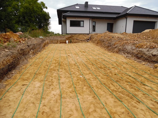 Gruntowy wymiennik ciepła ułożony w wykopie przed budynkiem mieszkalnym, widoczne zielone rury w piasku.