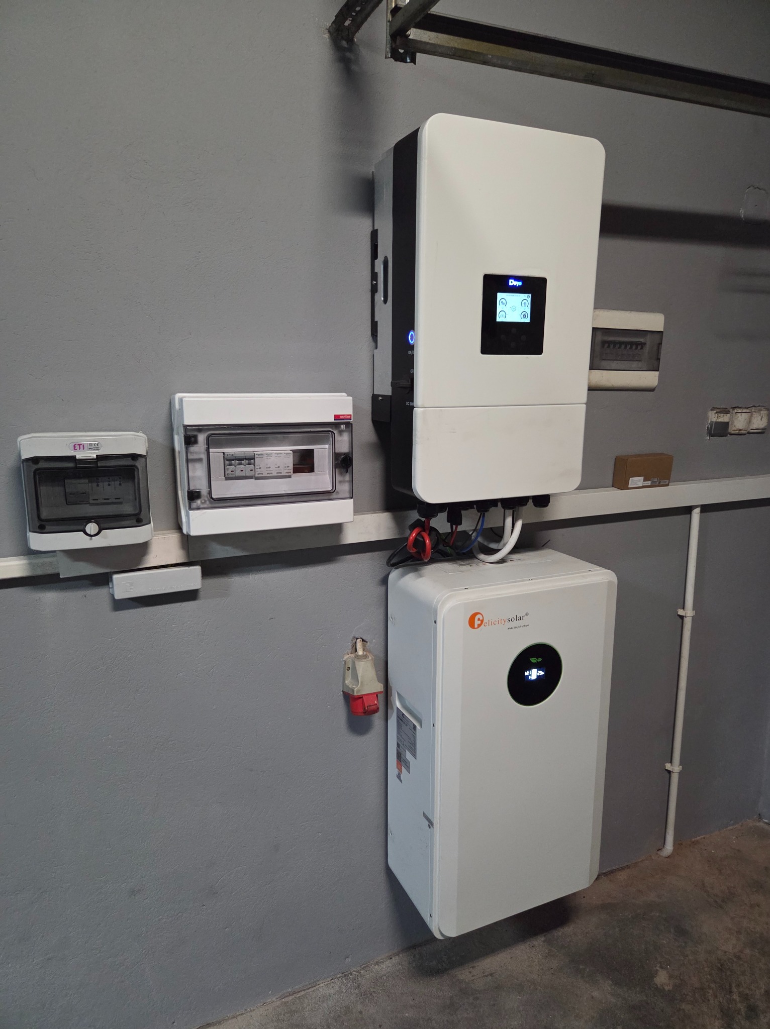 Biały inwerter fotowoltaiczny Deye i magazyn energii Felicity Solar zamontowane na szarej ścianie wewnątrz budynku. Widoczne skrzynki elektryczne i gniazdo siłowe.
