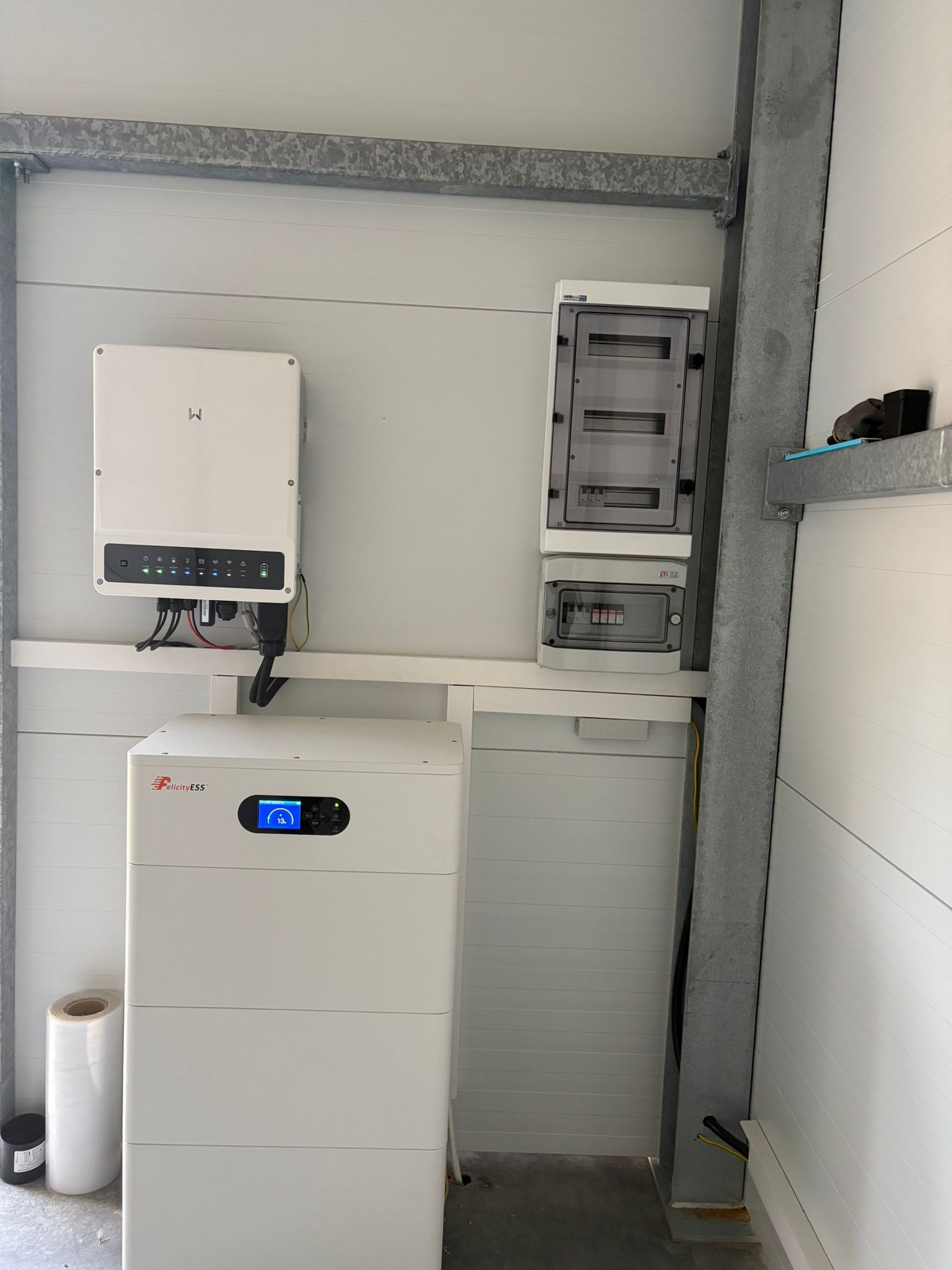 Biały magazyn energii Felicity ESS z inwerterem i rozdzielnicą na ścianie z paneli. Widoczne kable i przewody. Instalacja w pomieszczeniu z metalową konstrukcją.