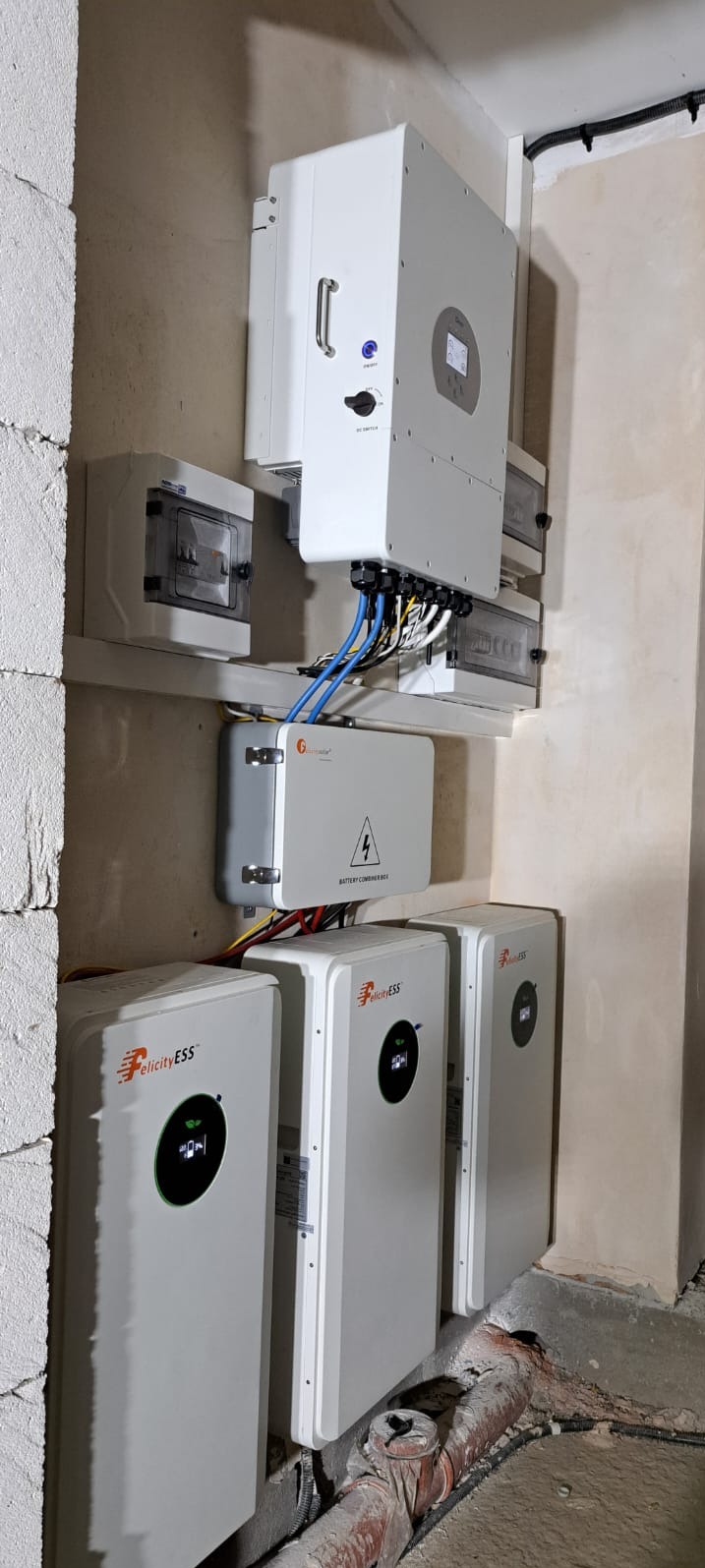 Instalacja magazynów energii Felicity ESS z podłączonym inwerterem i skrzynką bezpiecznikową na ścianie z surowego betonu. Widoczne kable i rury.