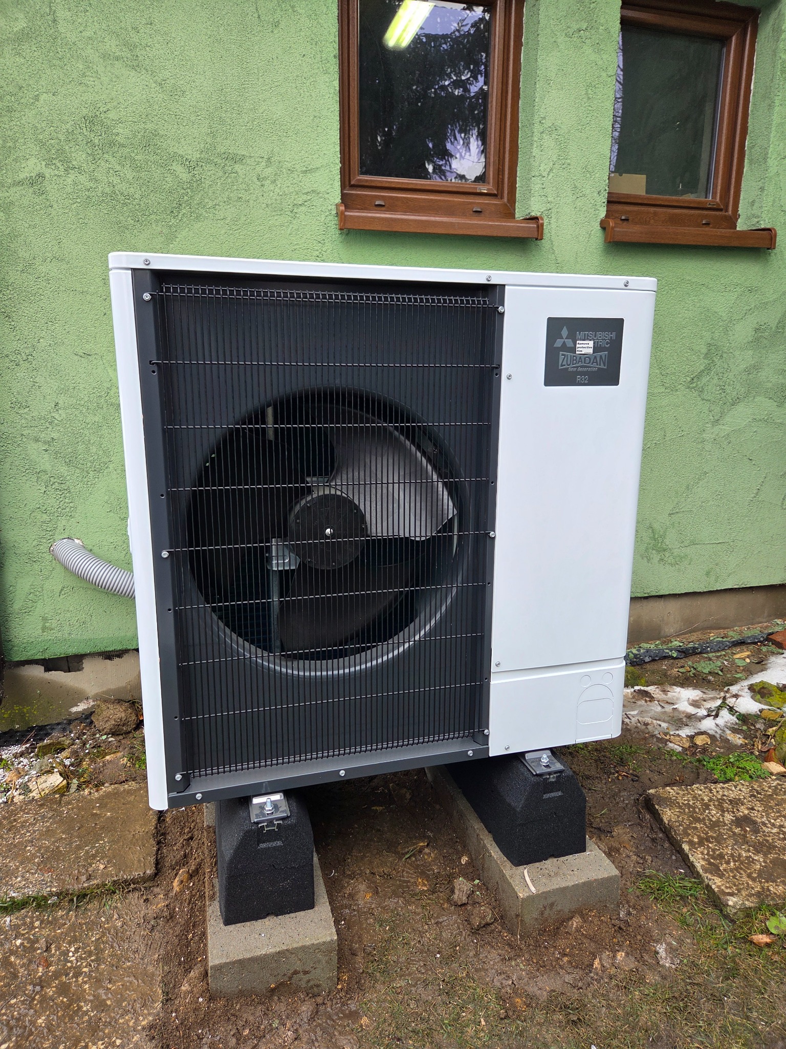 Biała pompa ciepła Mitsubishi Zubadan R32, zamontowana na betonowych podstawach przy zielonej ścianie budynku z dwoma oknami. Widoczny szary przewód i fragment trawnika.