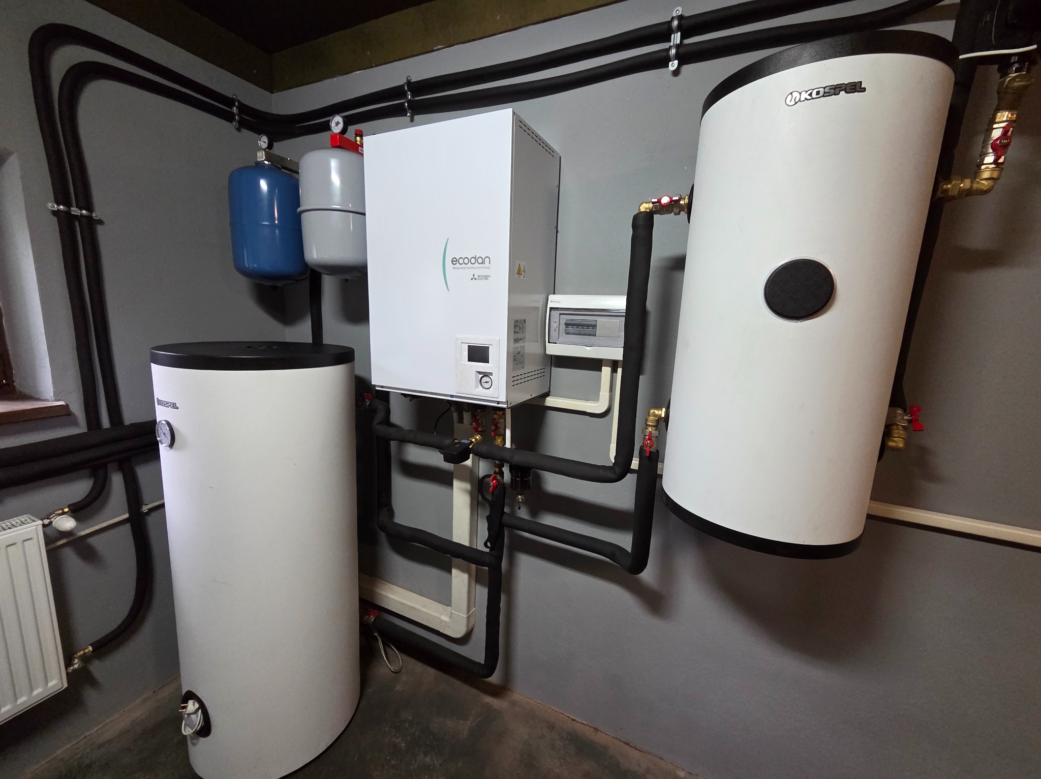 Zestaw urządzeń grzewczych: pompa ciepła Ecodan, dwa zbiorniki Kospel, instalacja rurowa w pomieszczeniu technicznym. Widoczne zawory i manometry.