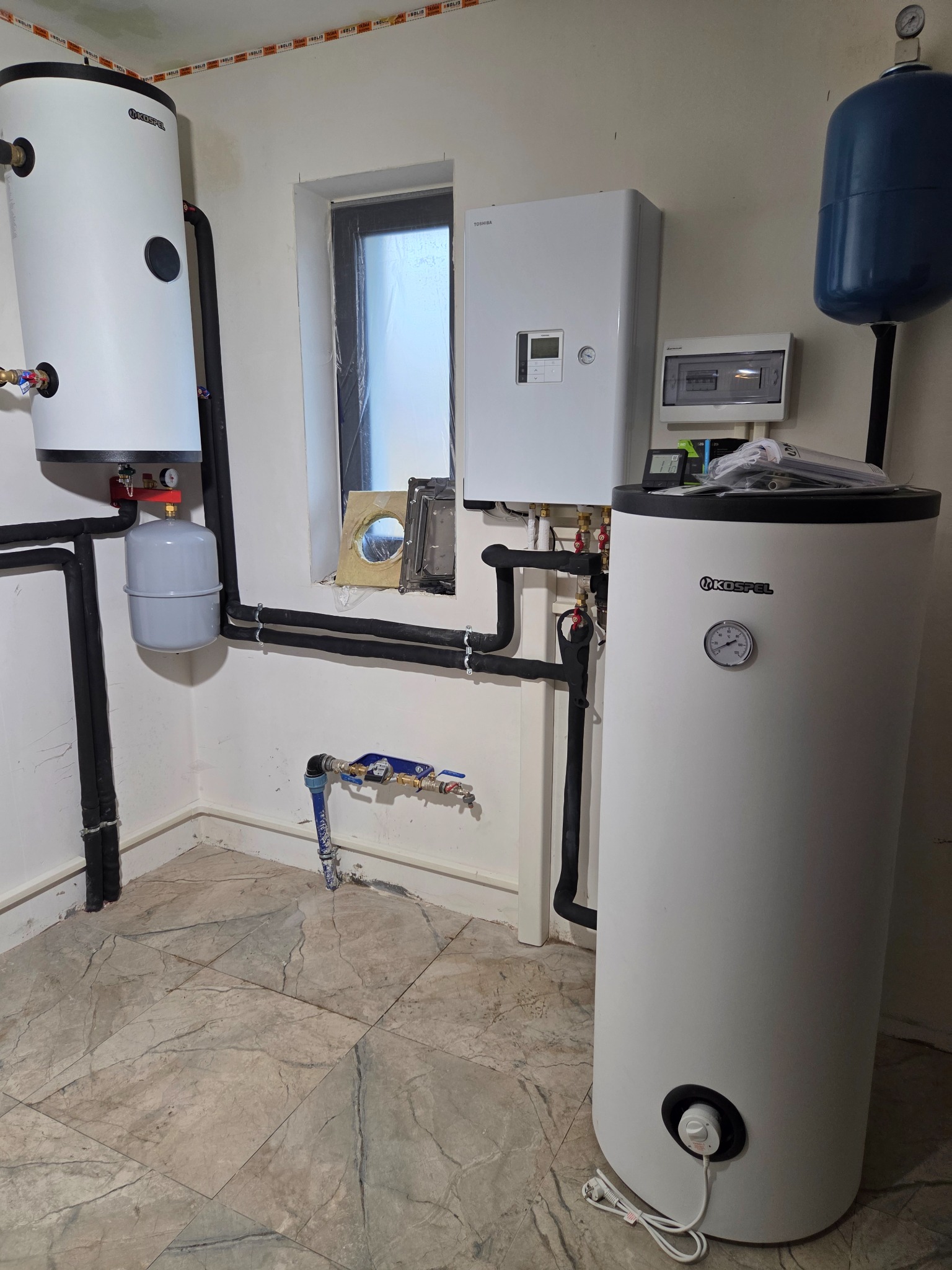 Biały zasobnik ciepłej wody Kospel z czarnymi rurami w kotłowni, obok biały piec Toshiba i niebieski zbiornik wyrównawczy. Widoczne przyłącza wodne i instalacja elektryczna.