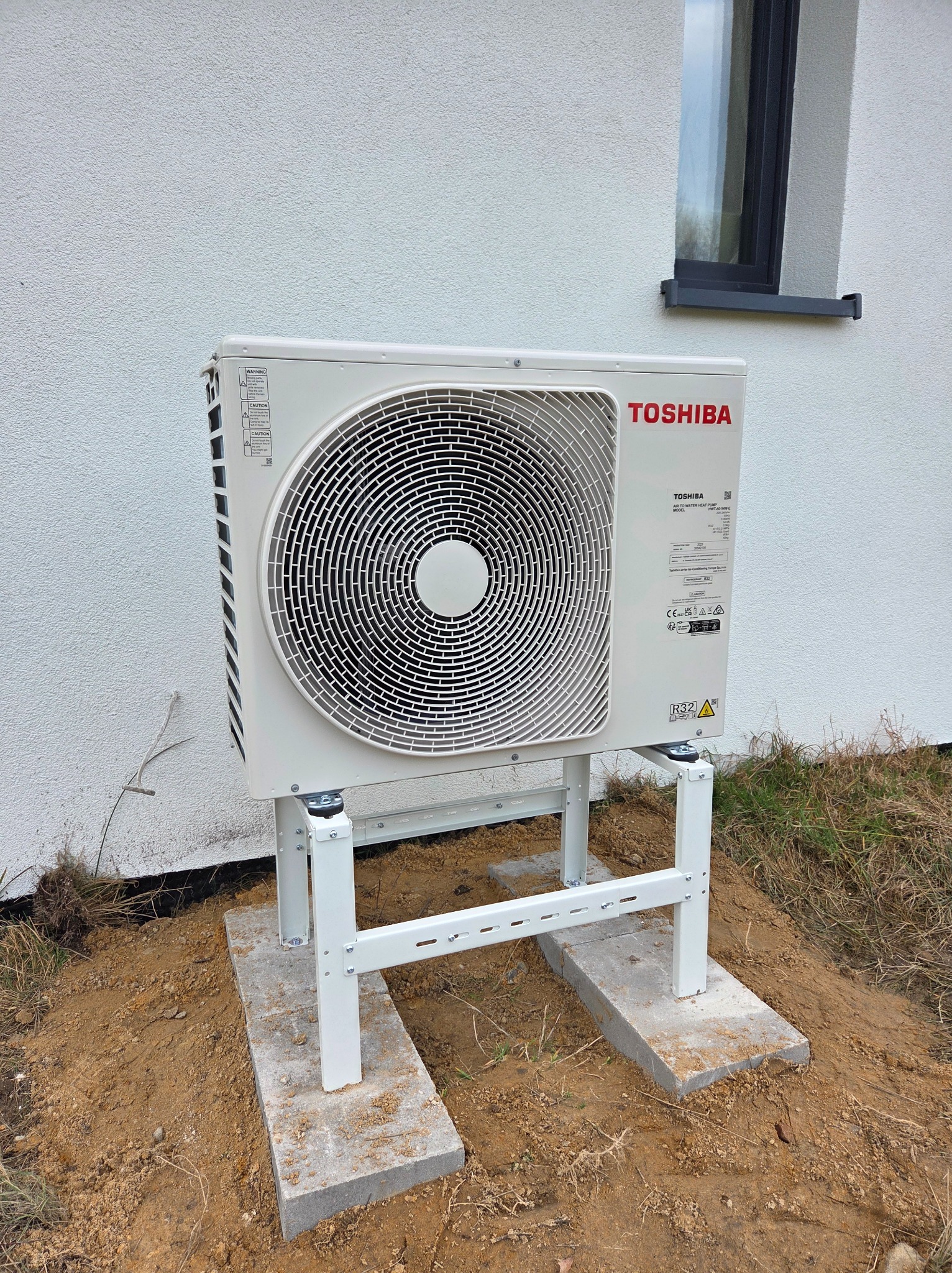 Biała pompa ciepła Toshiba na metalowym stelażu, ustawiona na betonowych płytach przed jasnym budynkiem z małym oknem. Widoczne elementy instalacji i otoczenie.