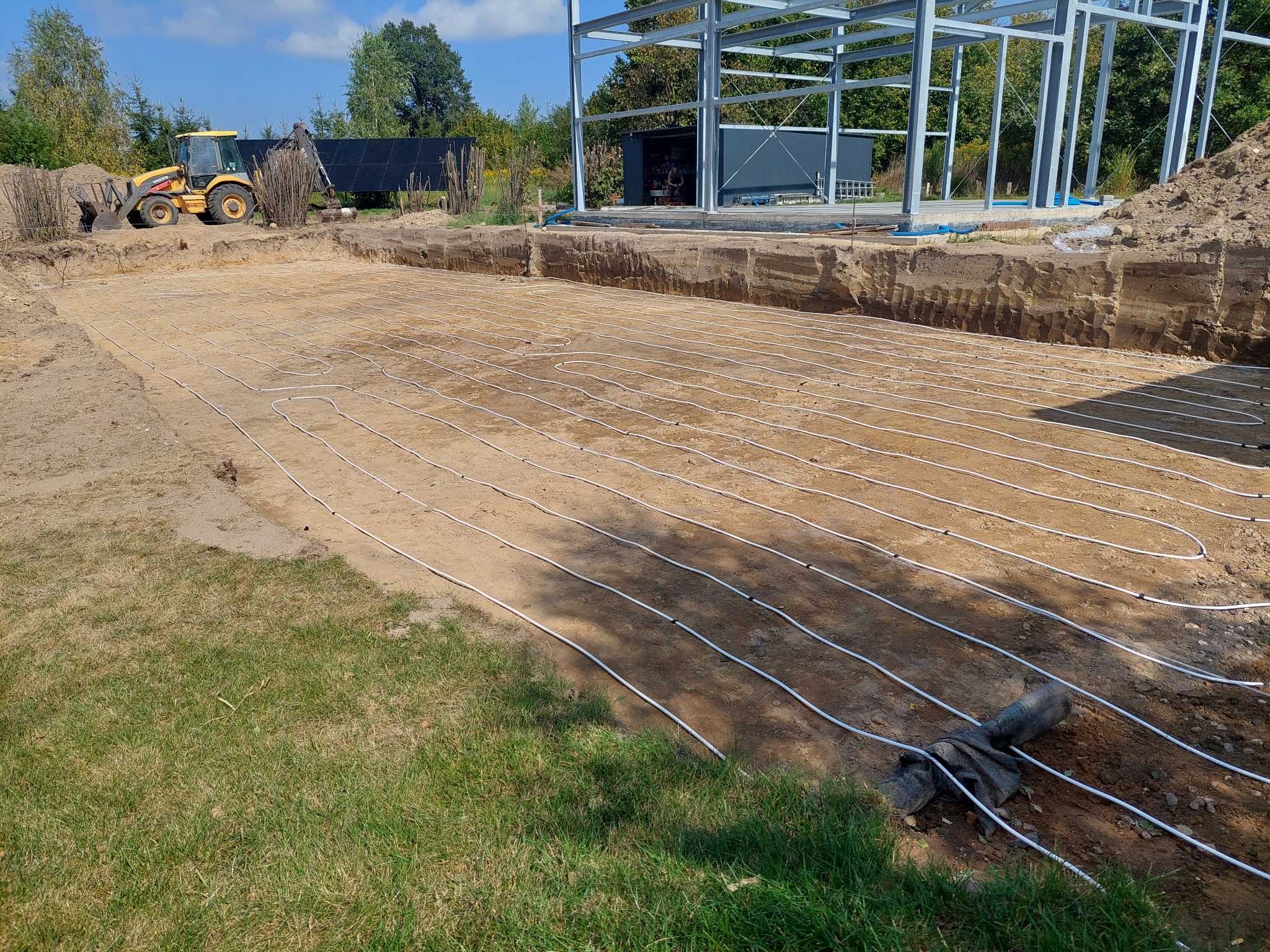 Instalacja ogrzewania podłogowego w gruncie pod budynek, widoczne białe rury ułożone w wykopie, w tle konstrukcja stalowa budynku i koparka.