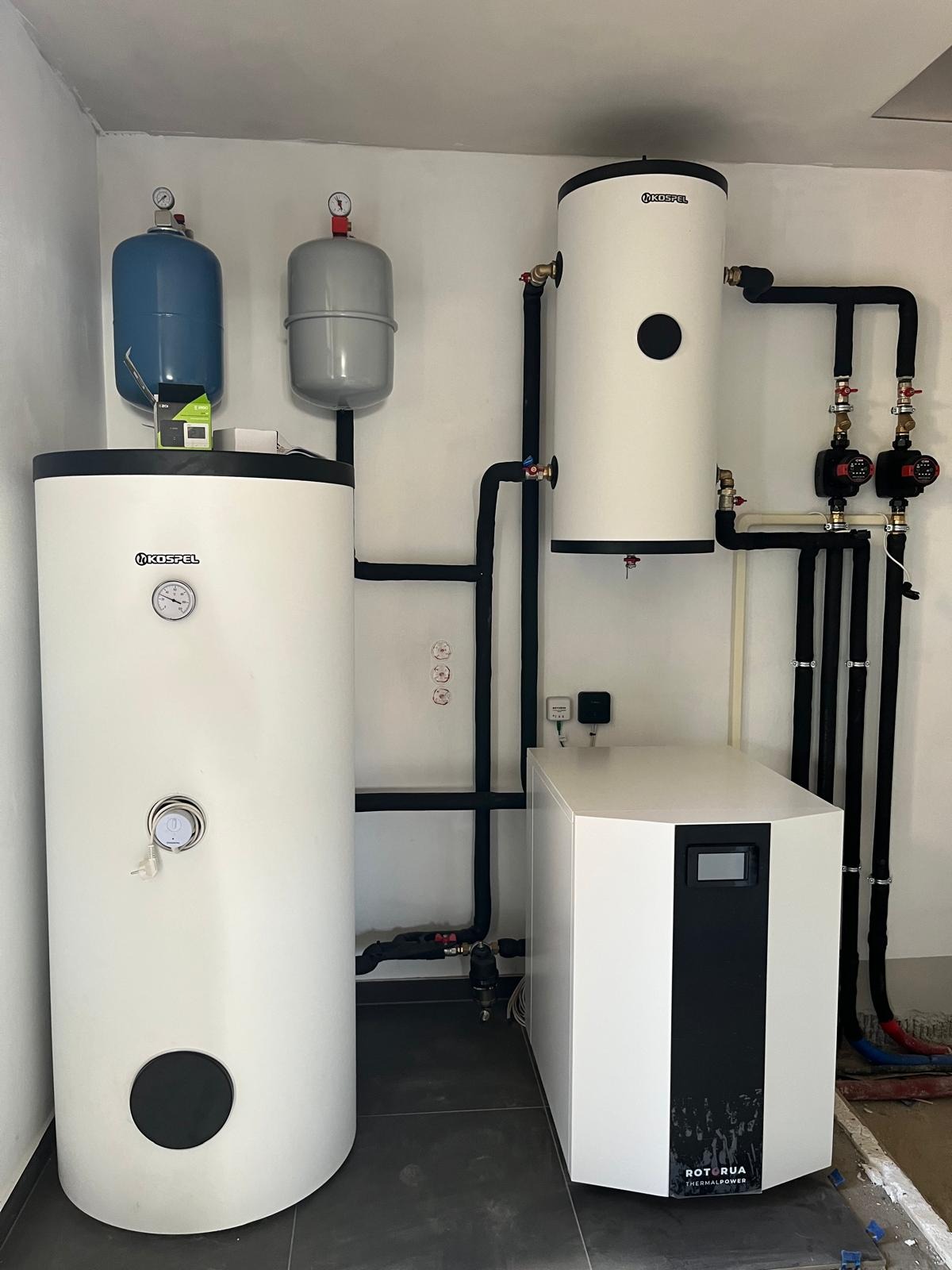 Kompletna instalacja kotłowni z białym zbiornikiem Kospel, szarym i niebieskim naczyniem wzbiorczym oraz białą pompą ciepła Rotorua Thermalpower. Czarna armatura.