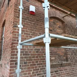 Czerwono-biała zewnętrzna syrena alarmowa zamontowana na ceglanej ścianie budynku, widoczna zza metalowych rusztowań.