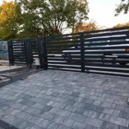 Nowoczesna, pozioma brama wjazdowa z automatyką, szara kostka brukowa przed posesją, w tle widoczny samochód i maszyna do betonu.