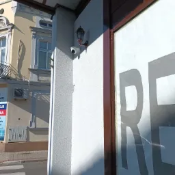 Zewnętrzna kamera IP zamontowana na białej ścianie budynku obok stylizowanej lampy, widoczny fragment elewacji i przejście dla pieszych.