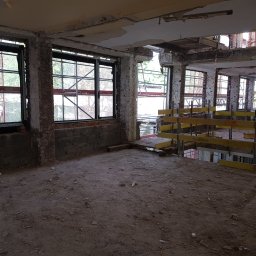 Yevhen Havrilov - Wnętrze budynku w trakcie remontu, widoczne stare okna z metalowymi ramami, fragmenty ceglanych ścian, uszkodzony sufit i podłoga pokryta gruzem. W tle widoczne rusztowania i dalsza część budynku...